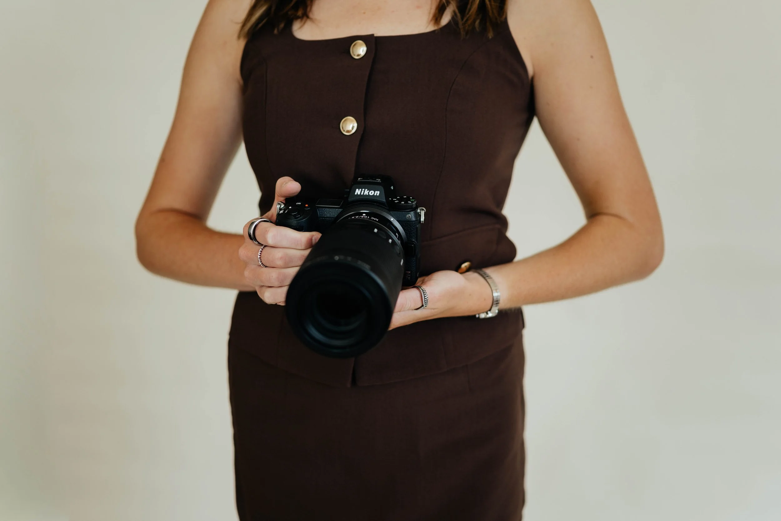 A woman in a brown sleeveless dress holding a Nikon camera with a large lens, standing against a plain light background.