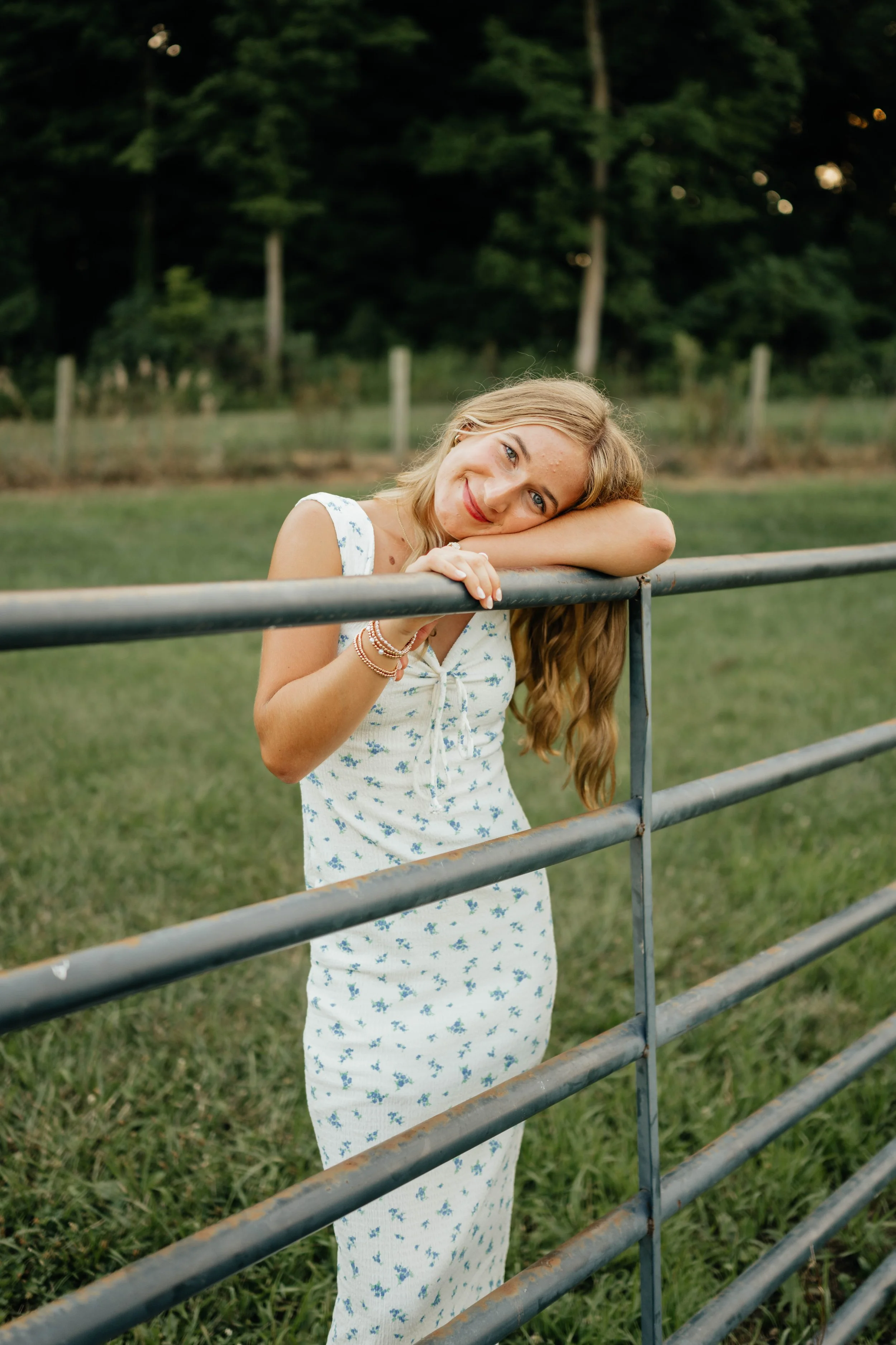 Young woman with long wavy hair leaning on a metal fence, smiling and looking at the camera in a grassy outdoor setting with trees in the background.