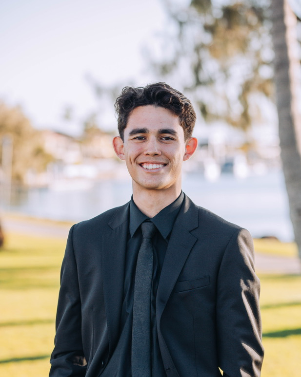 A young man in a black suit and tie smiling outdoors near a body of water with trees in the background.