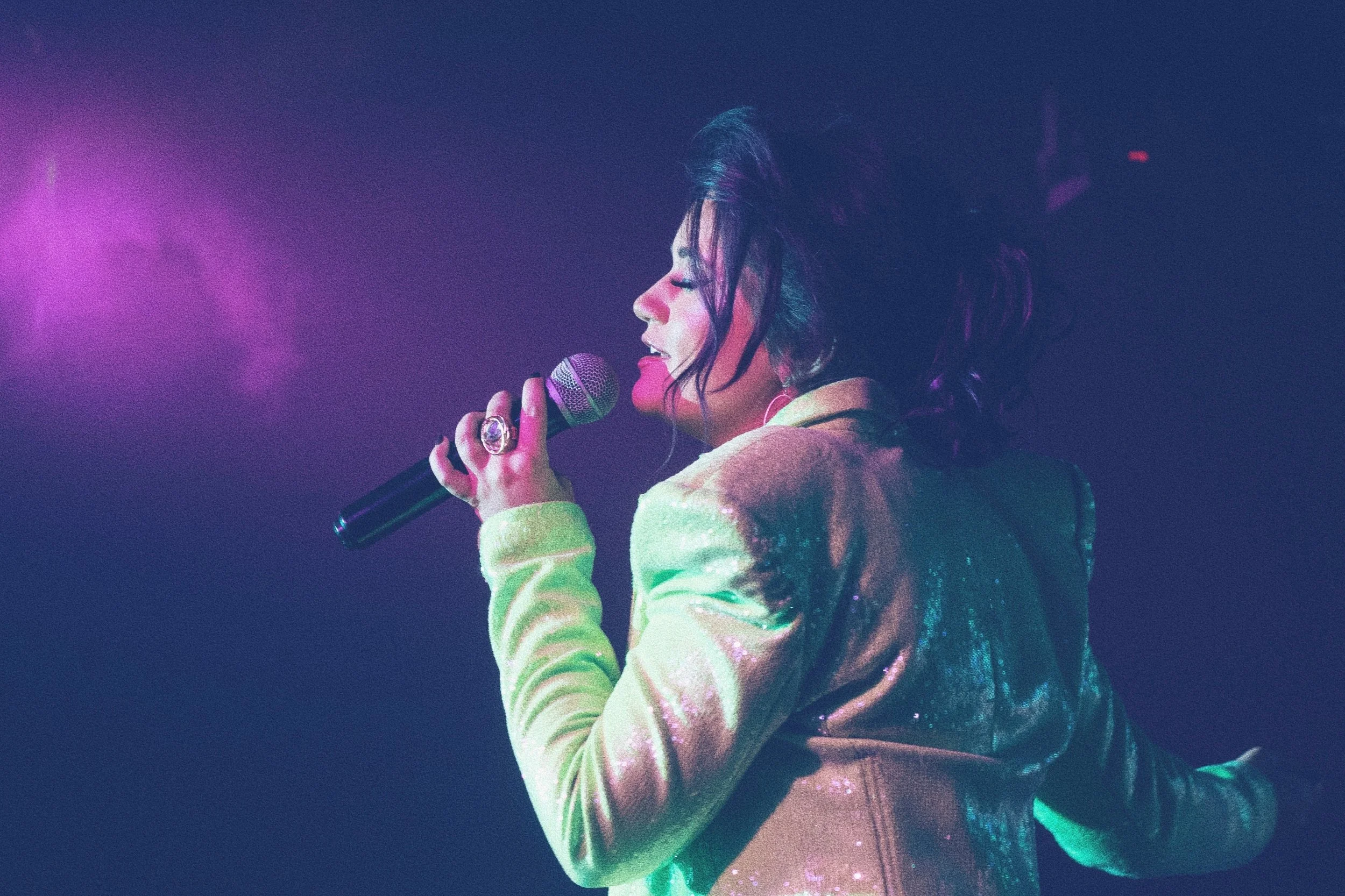 A woman singing into a microphone on stage, illuminated by colorful purple and green stage lights, with her hair styled in a messy, voluminous fashion and wearing an iridescent blazer.