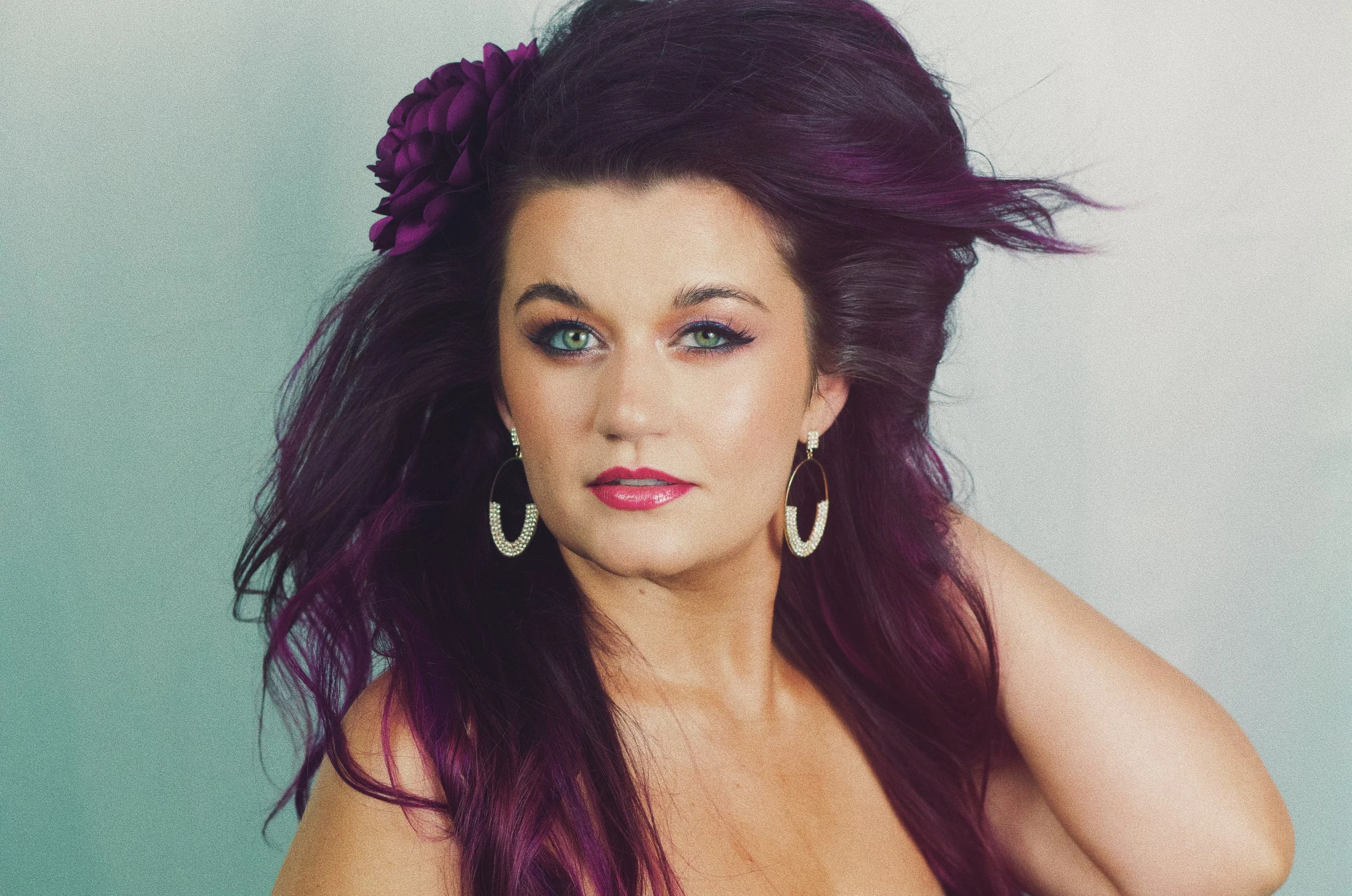 A woman with long, dark purple hair and bright green eyes, wearing large hoop earrings with rhinestones and pink lipstick, poses with her hand near her head and a purple flower in her hair.