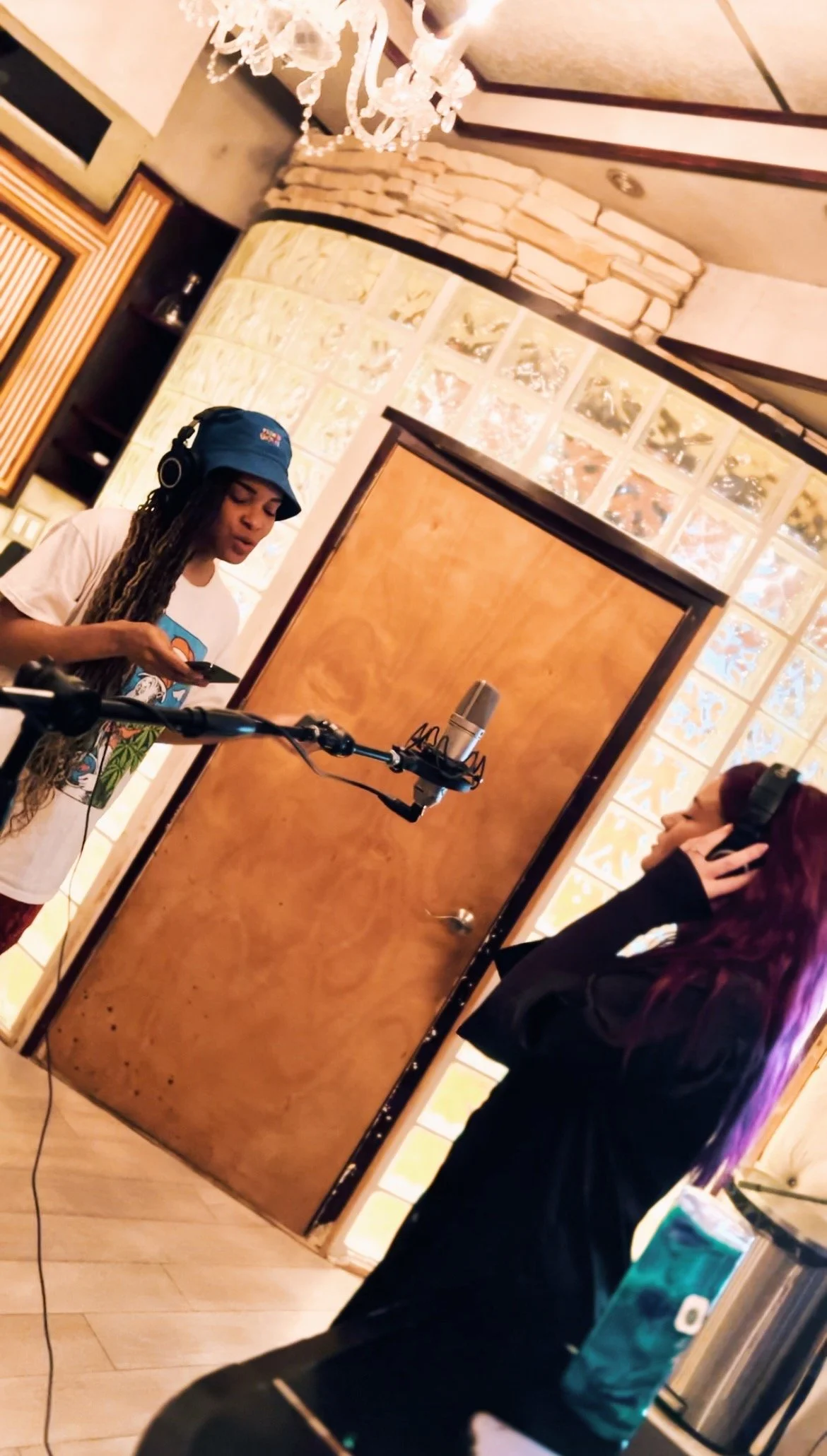 Two women in a recording studio, one with headphones and the other with a microphone, both wearing headphones and standing near a door with glass blocks in the background, with a chandelier hanging from the ceiling.