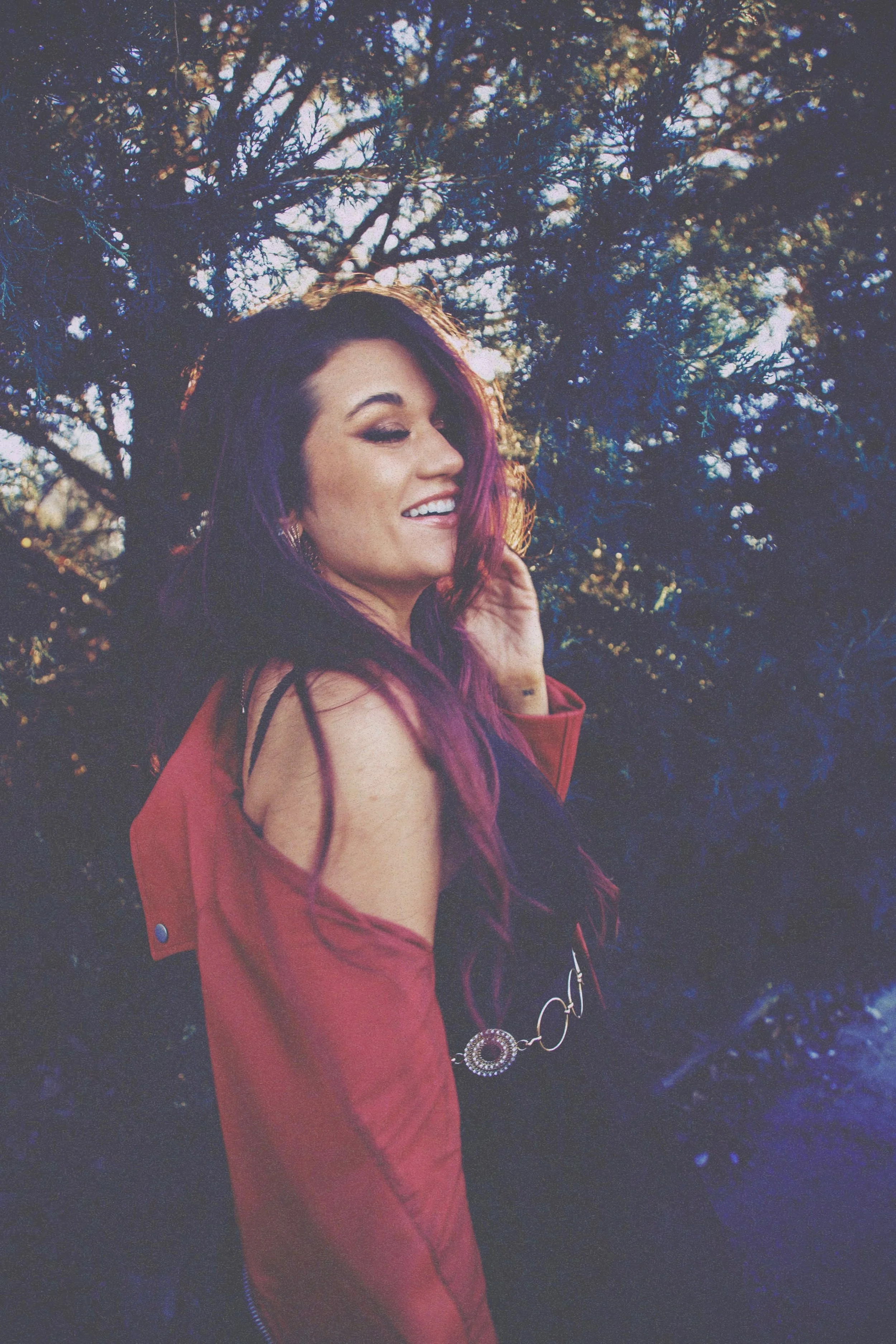 A woman with purple hair and makeup, smiling and touching her hair, standing outdoors in front of a tree during sunset, wearing a black top and a red jacket.