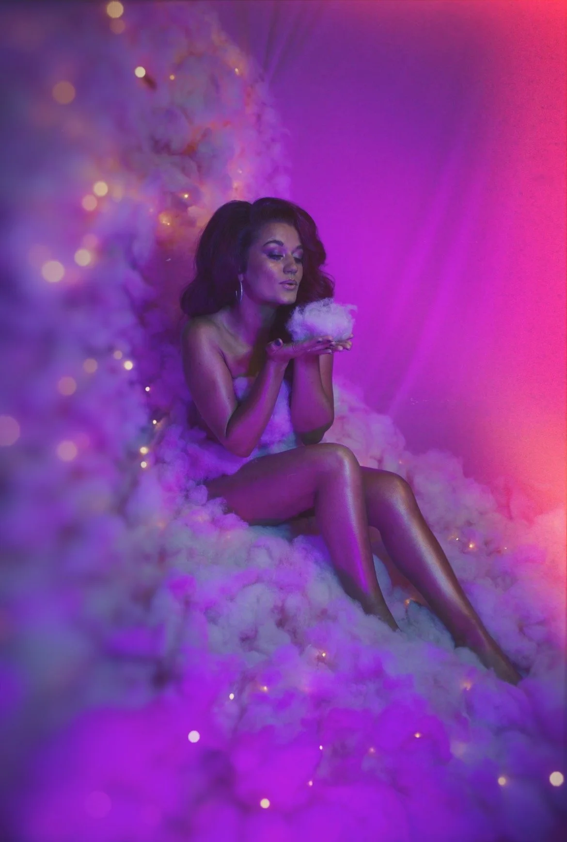 A woman with dark hair and hoop earrings is sitting on fluffy clouds, holding a handful of cotton candy, with purple and pink lighting creating a dreamy atmosphere.