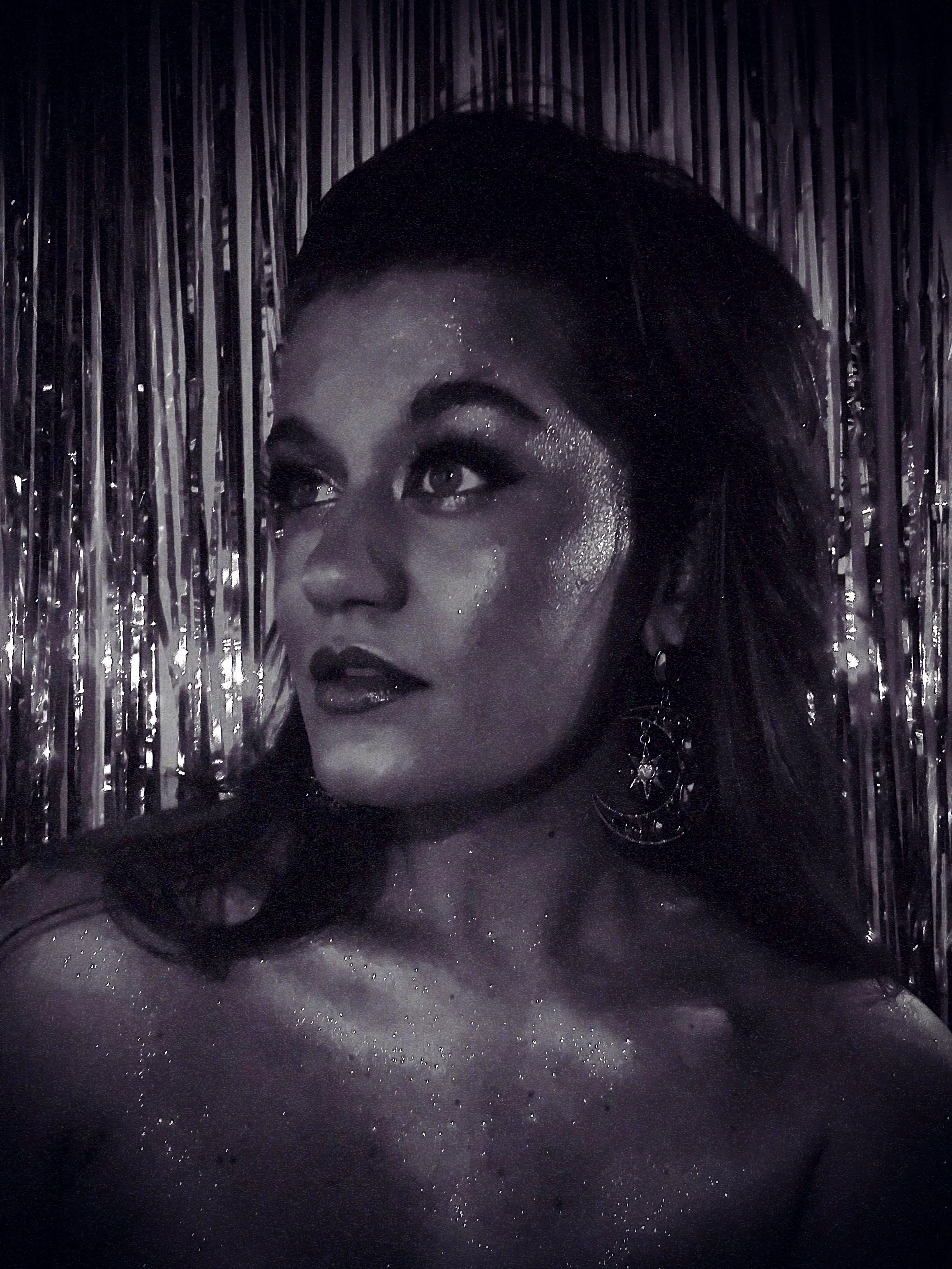 A woman with glitter on her face and shoulders, posing against metallic streamers, wearing earrings with star and moon designs.