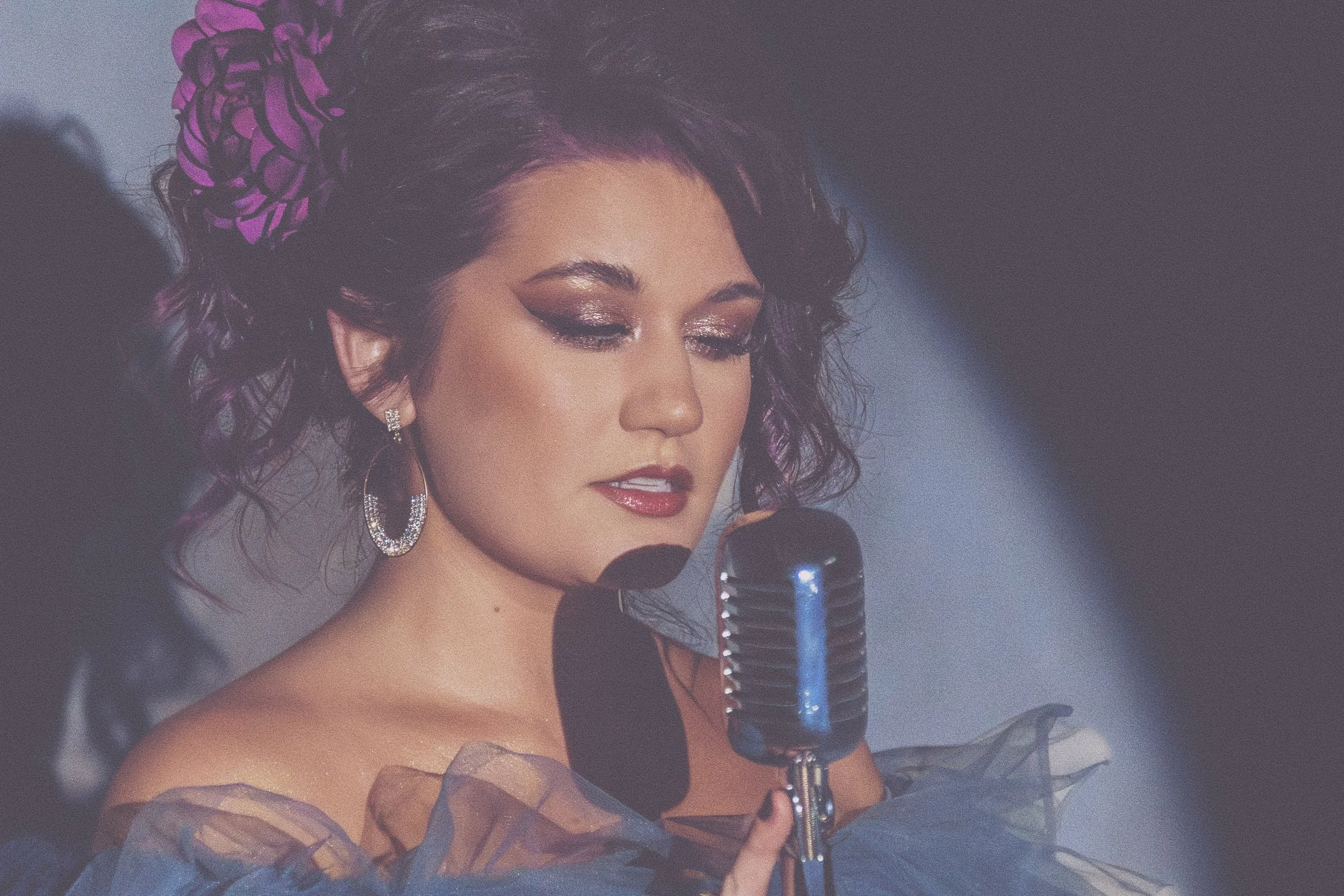 A woman with curly dark hair and purple floral hair accessory singing into a vintage microphone, wearing shimmer makeup and earrings.