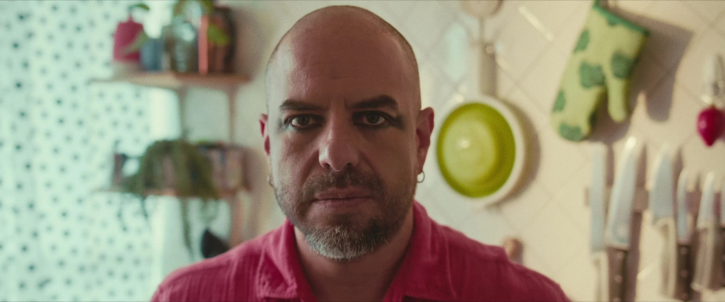 A bald man with a beard and earrings wearing a red shirt looking into the camera in a kitchen with hanging utensils and a green plate in the background.
