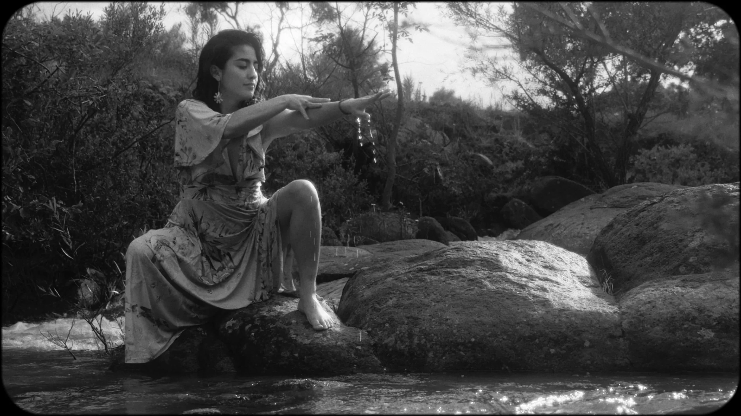 Black and white photo of a woman sitting on rocks in a stream, with her arms extended forward, holding a small object, surrounded by trees and nature.