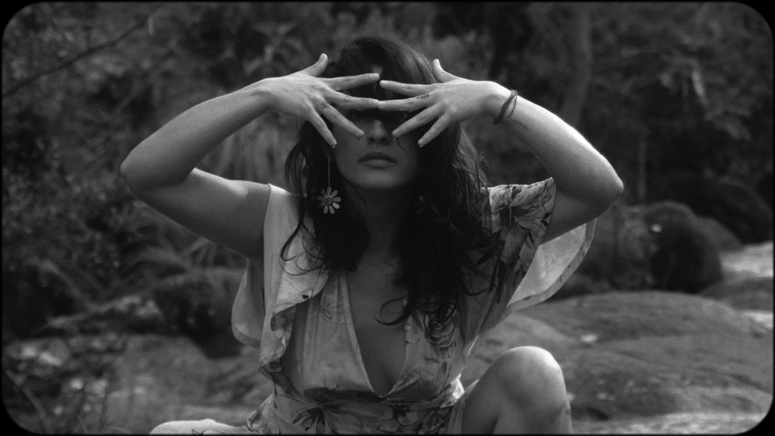 A woman with dark, wavy hair wearing floral clothing and an earring with a daisy flower design, posing outdoors with her hands covering her eyes.