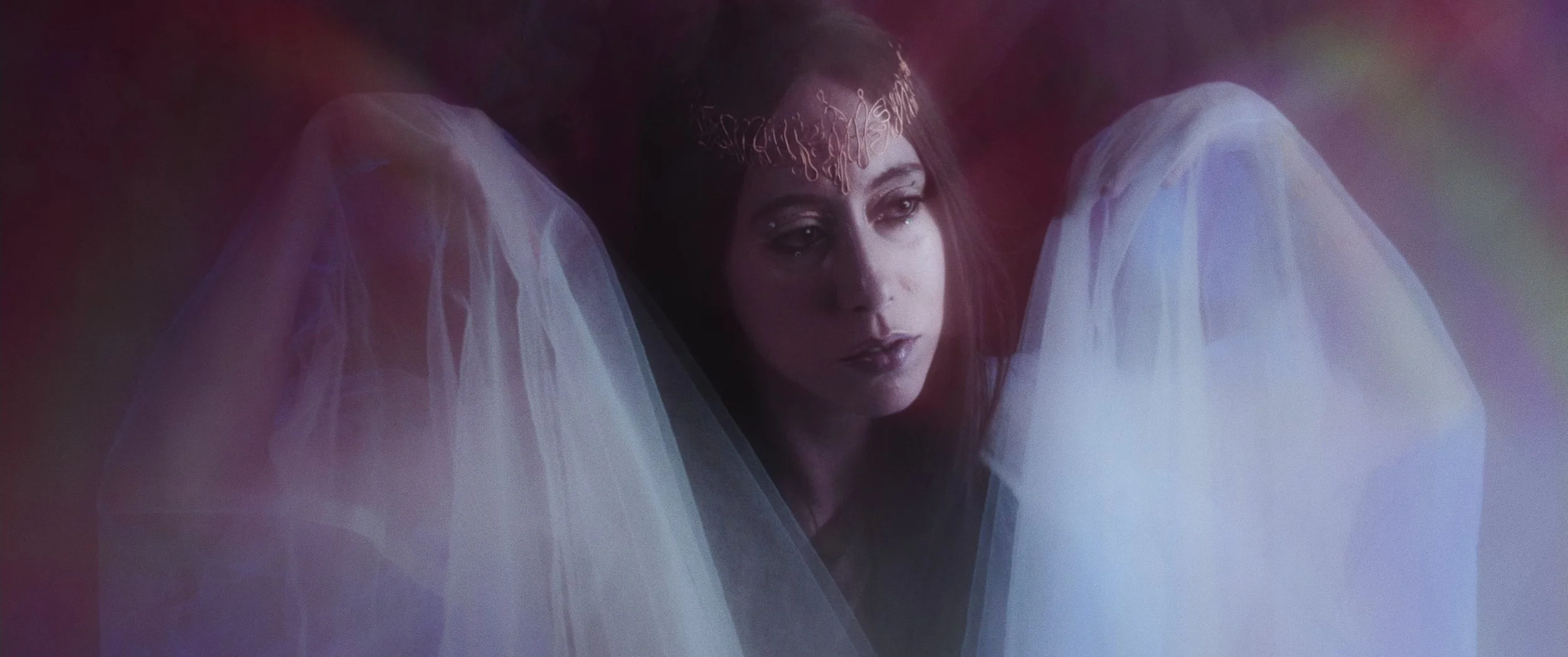A woman with dark hair and makeup, wearing a metallic headpiece, surrounded by white sheer fabric, with a dark background.