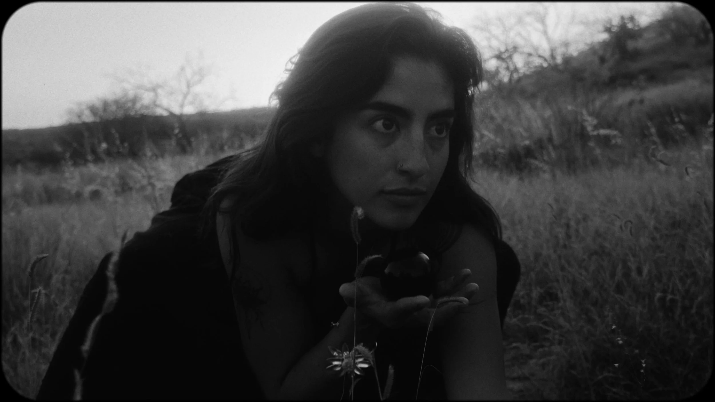 A woman with dark hair, holding a small object, is outdoors in a grassy field with trees in the background, captured in black-and-white.