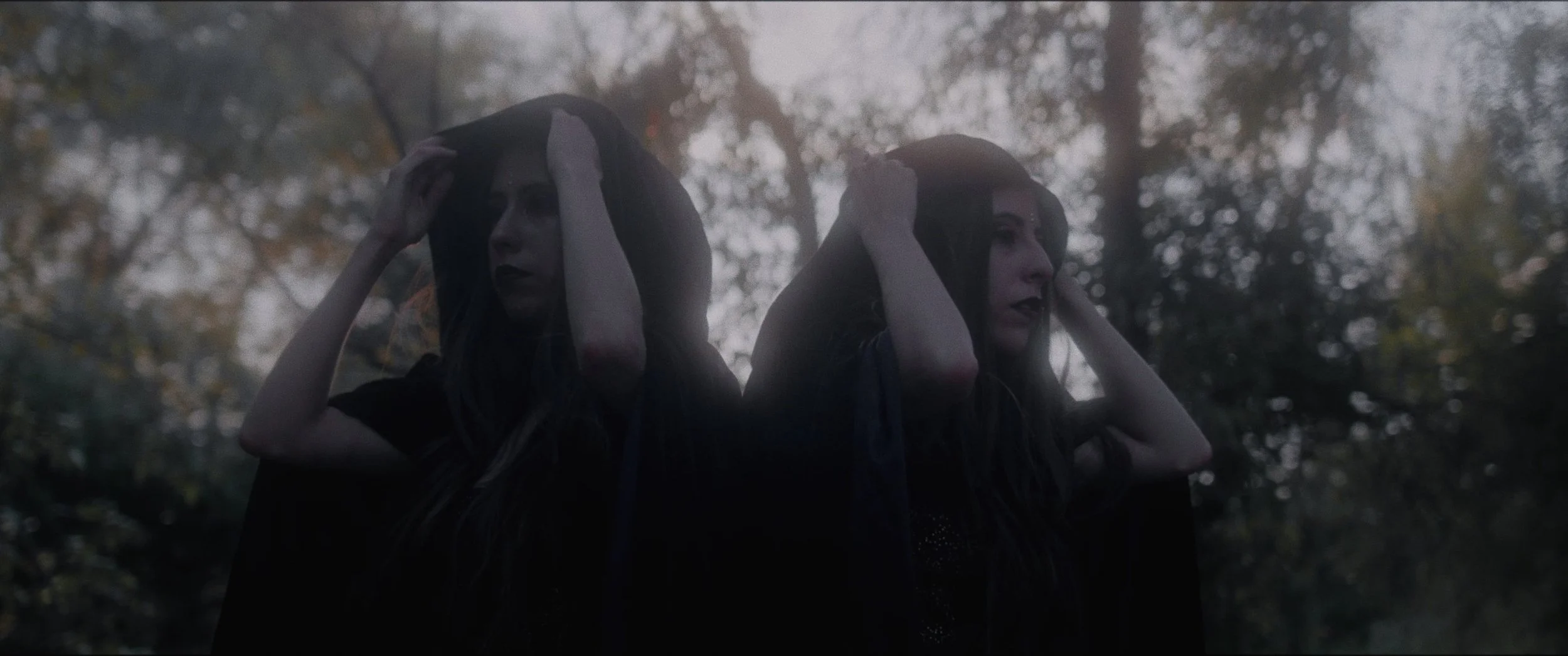 Two women with long dark hair wearing dark clothing, holding hoods over their heads, stand outdoors in a wooded area on a foggy day.