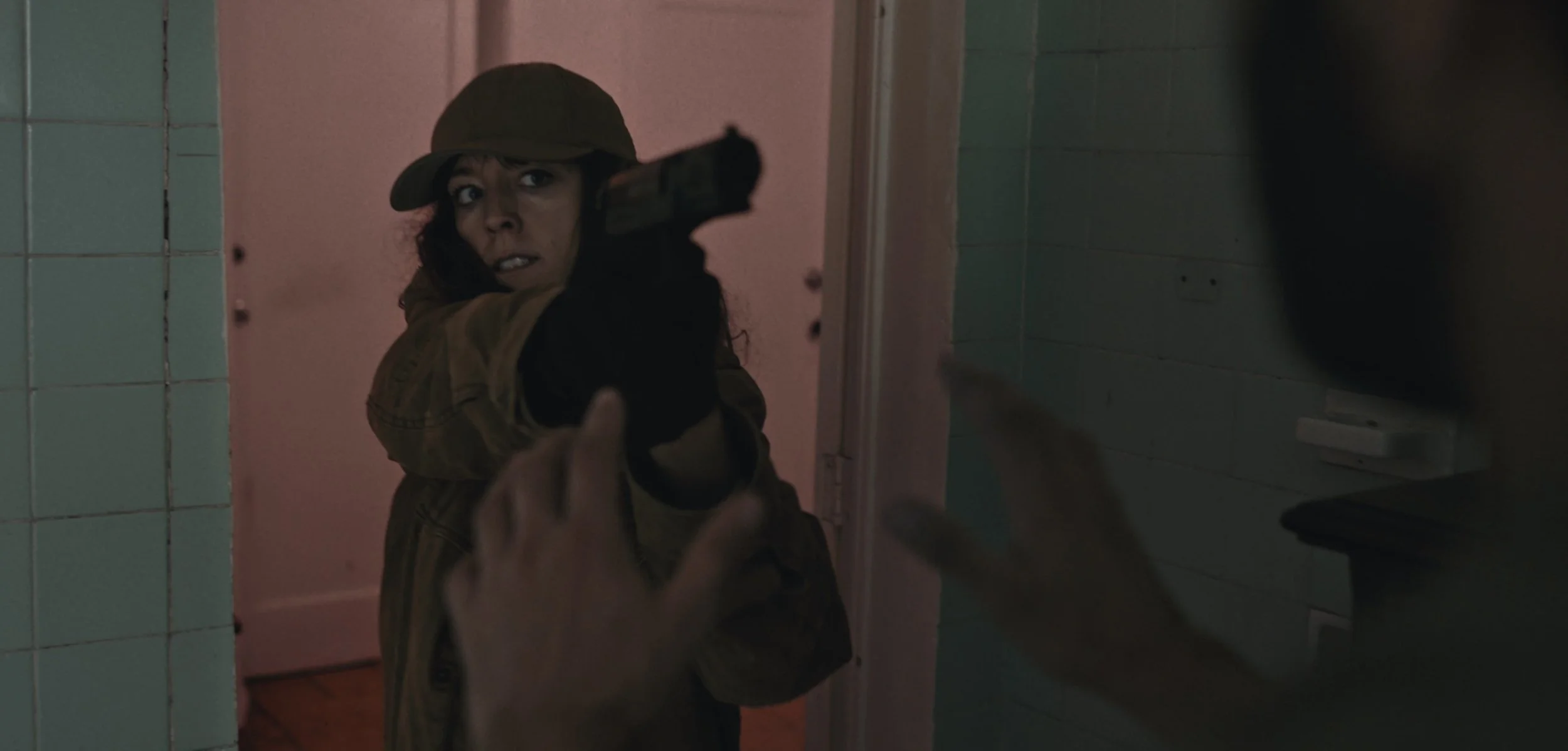 A woman in a military-style jacket and hat pointing a gun at someone outside the bathroom door, with a tense expression on her face.