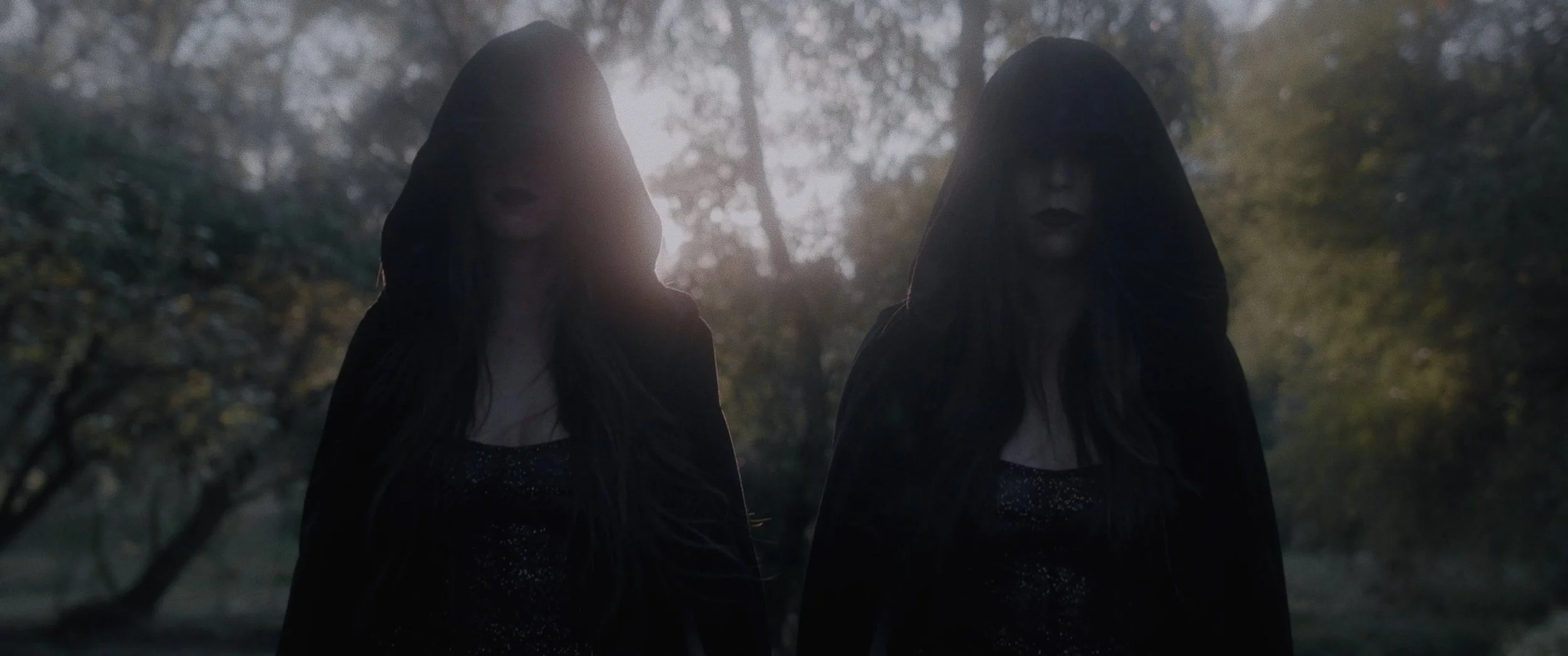Two women dressed in dark clothing with hoods over their heads standing outdoors in a wooded area during twilight, with sunlight filtering through the trees.