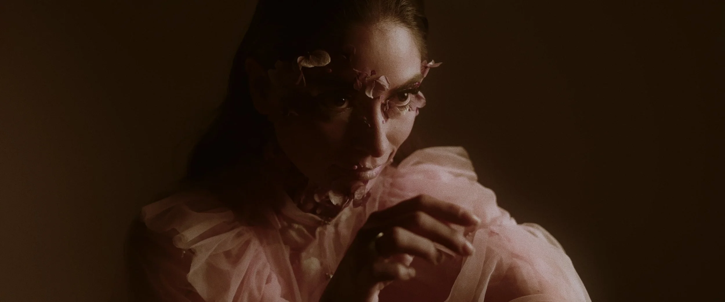 A woman with intense eyes covered partially by pink flower petals, wearing pink tulle clothing, in a dark, moody lighting setting.
