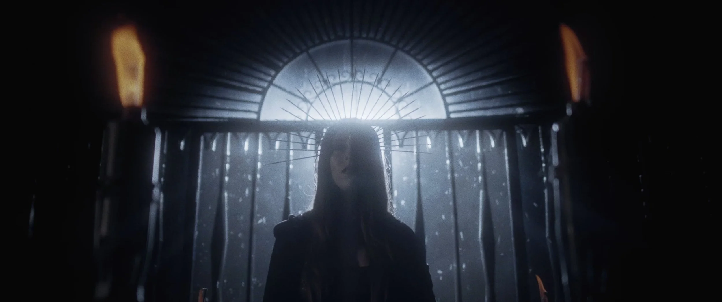 A woman standing behind a black wrought-iron gate with sunlight shining through, creating a dramatic backlit effect.