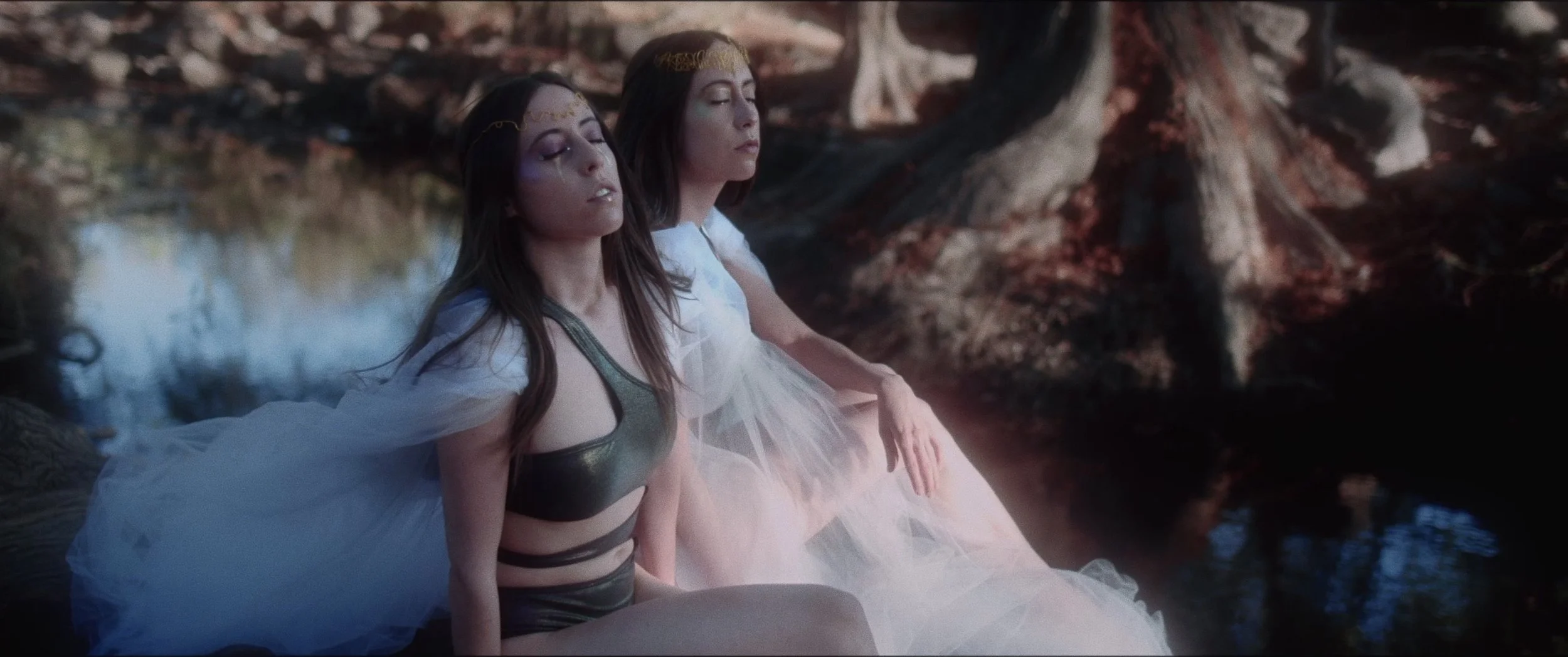 Two women with long hair and gold headbands, sitting by a river with their eyes closed, wearing white and dark clothing, surrounded by trees and rocks.