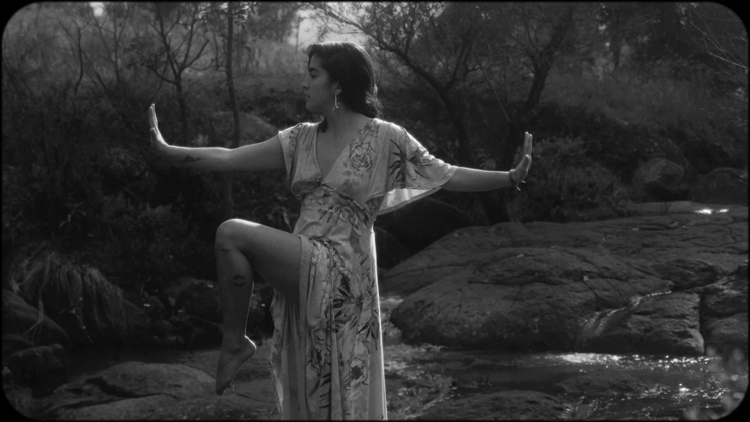 A woman in a floral dress balancing on one leg on rocks in a stream, with her arms extended in a yoga pose, in an outdoor natural setting with trees and water, in black and white.
