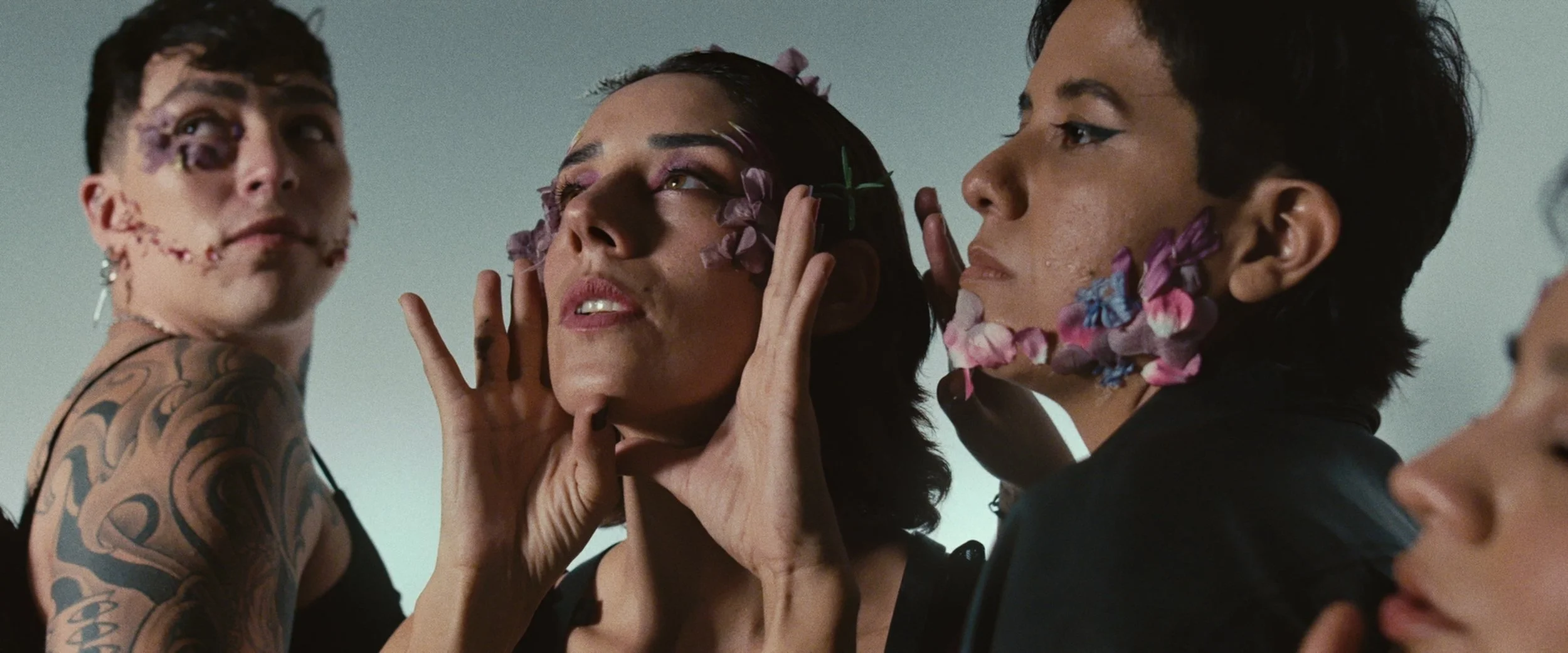 Three individuals, each with flowers on their faces, posing closely together. The person in the middle has their eyes closed and hands on their face, the person on the right has flowers on their cheek, and the person on the left has tattoos on their 