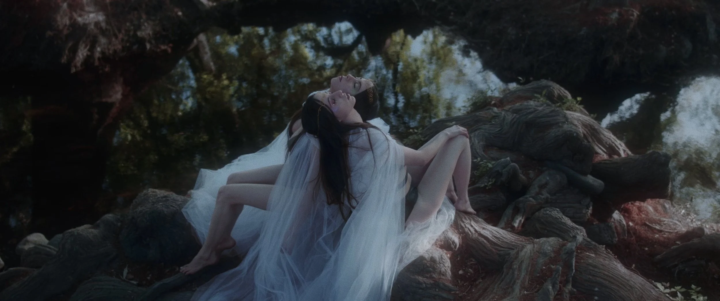 Two women dressed in white, lying on a large fallen tree by water, with their heads tilted back and eyes closed, surrounded by a natural outdoor setting.