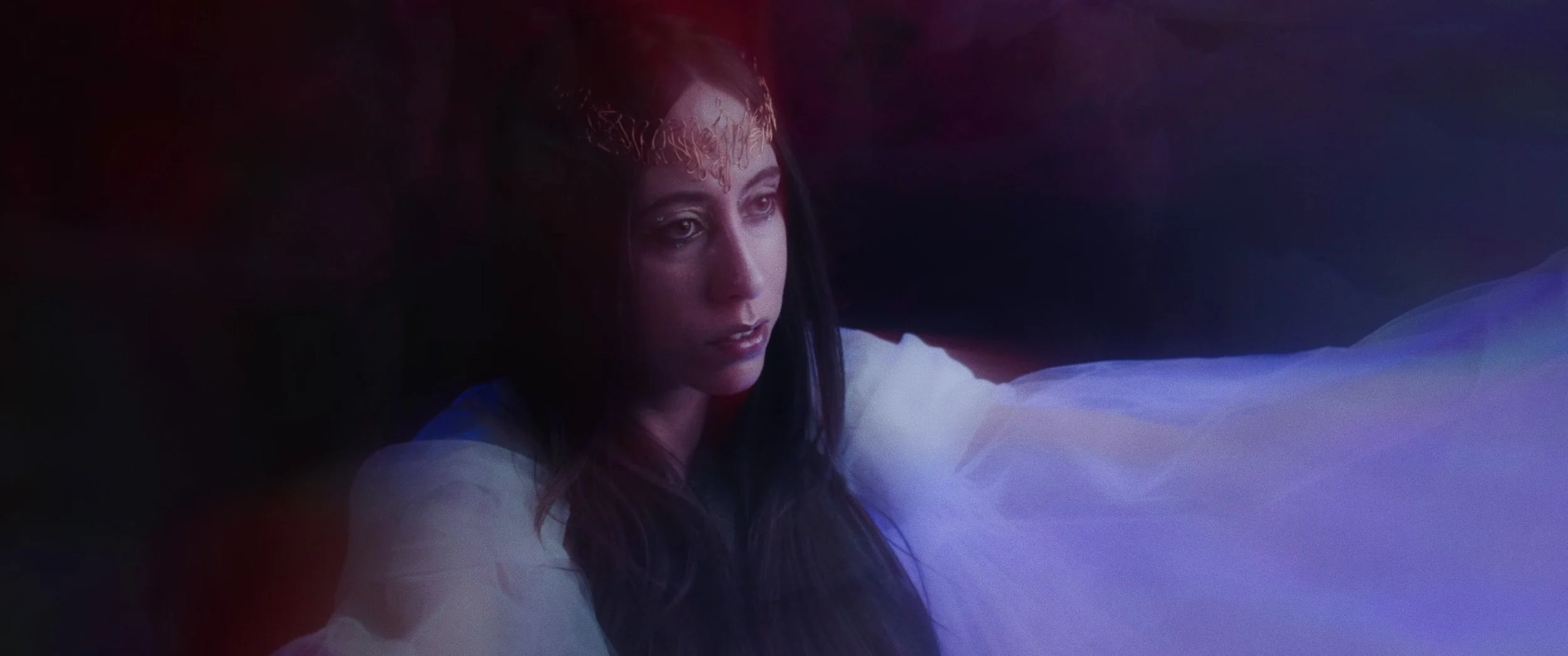A woman with long dark hair, wearing a crown, gazes thoughtfully in a dimly lit environment with purple and red lighting.