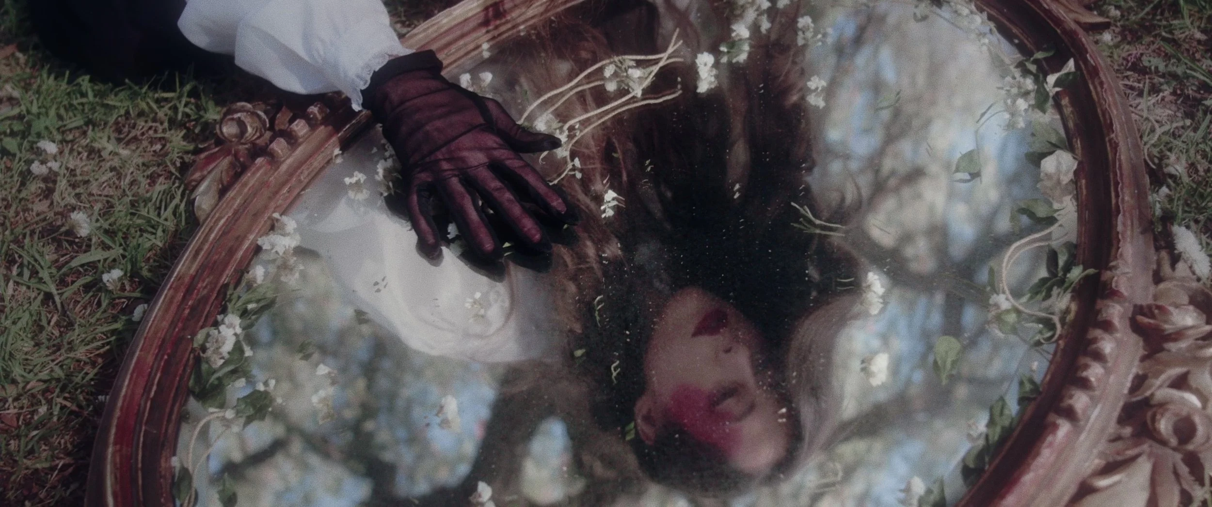 A person with dark hair and red lipstick rests their head on a vintage mirror with an ornate frame, lying on grass outdoors. The mirror reflects trees and sky.
