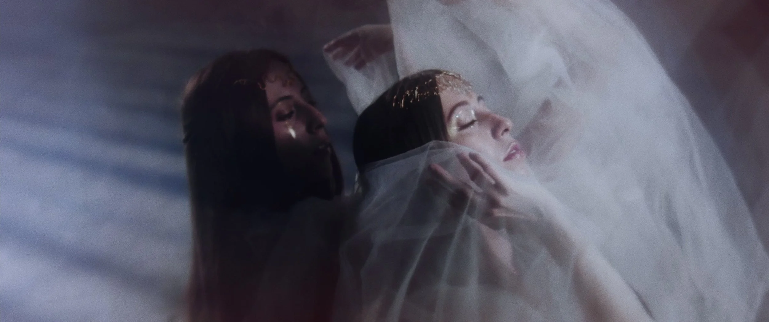 A woman lying down with eyes closed, covered with tulle fabric, with another woman behind her gently holding her face, in a soft, ethereal setting.