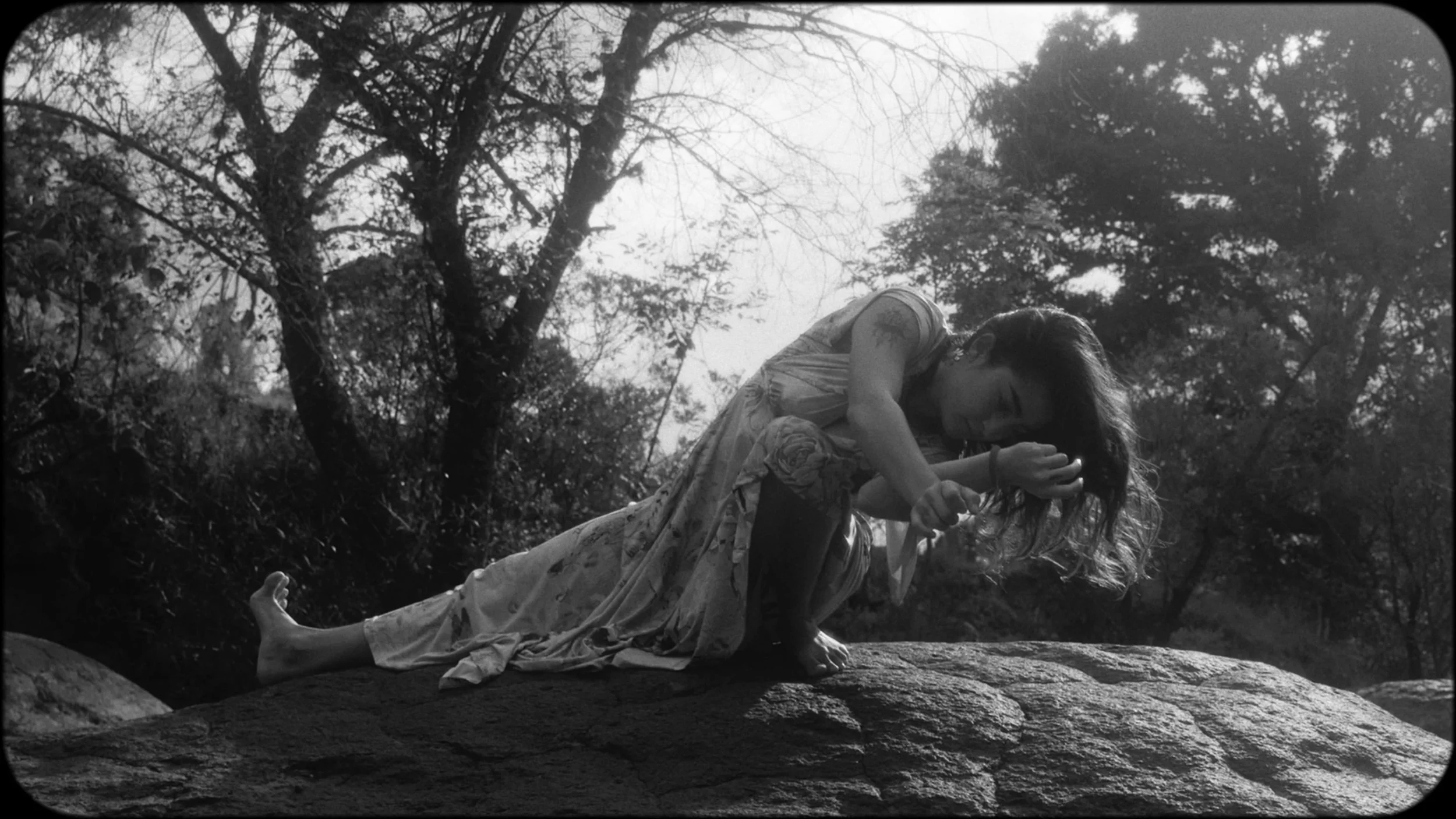 A woman dressed in a patterned dress crouches on a large rock outdoors, holding her hair with one hand, surrounded by trees and foliage in black and white.