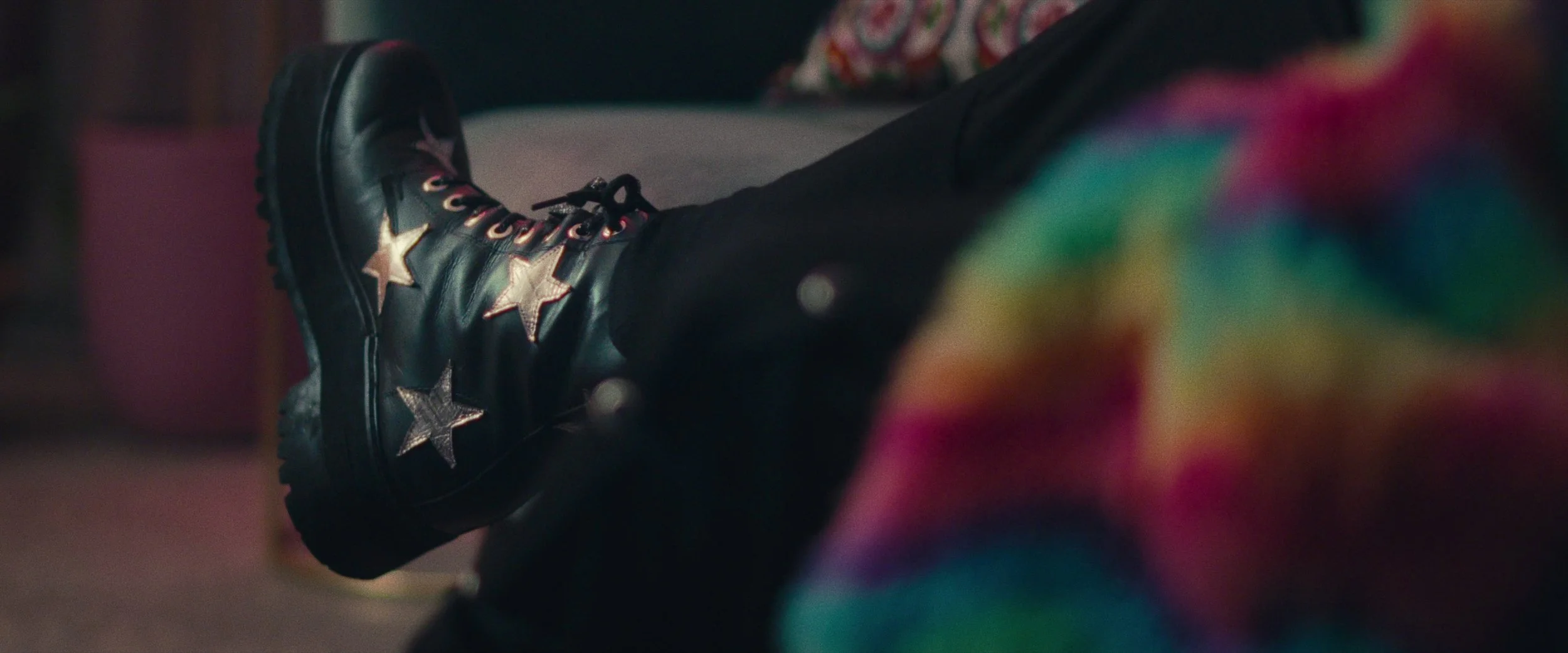 A person wearing black boots with gold and silver star designs, sitting with legs crossed. The person is also wearing rainbow-colored clothing seen in the foreground.
