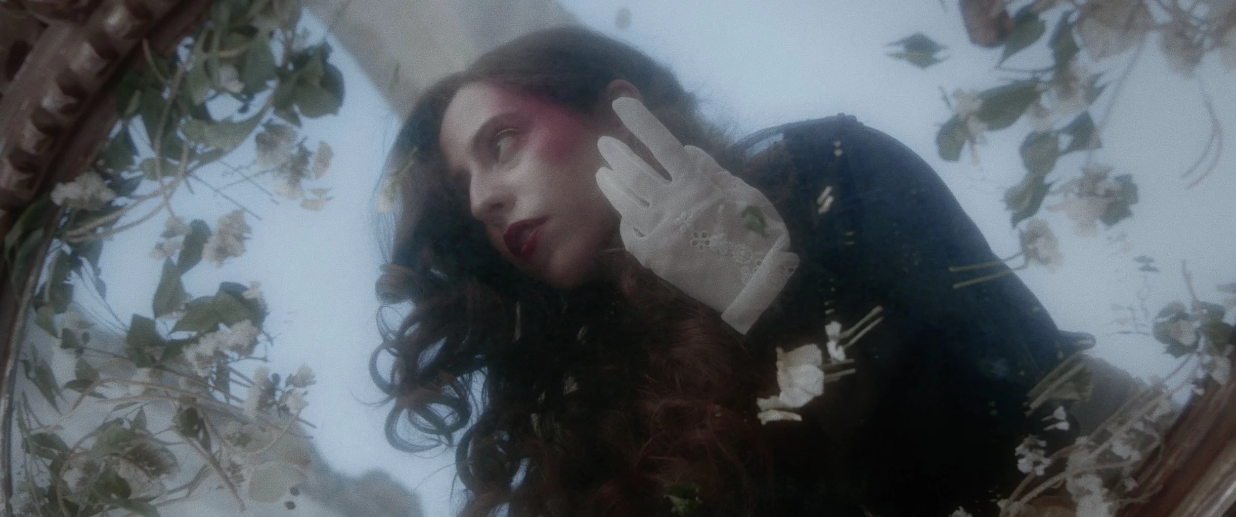 A woman with wavy dark hair and red makeup looking into a mirror, wearing a lace glove and dark clothing, with a cloudy or smoky background and dried flowers framing the mirror.