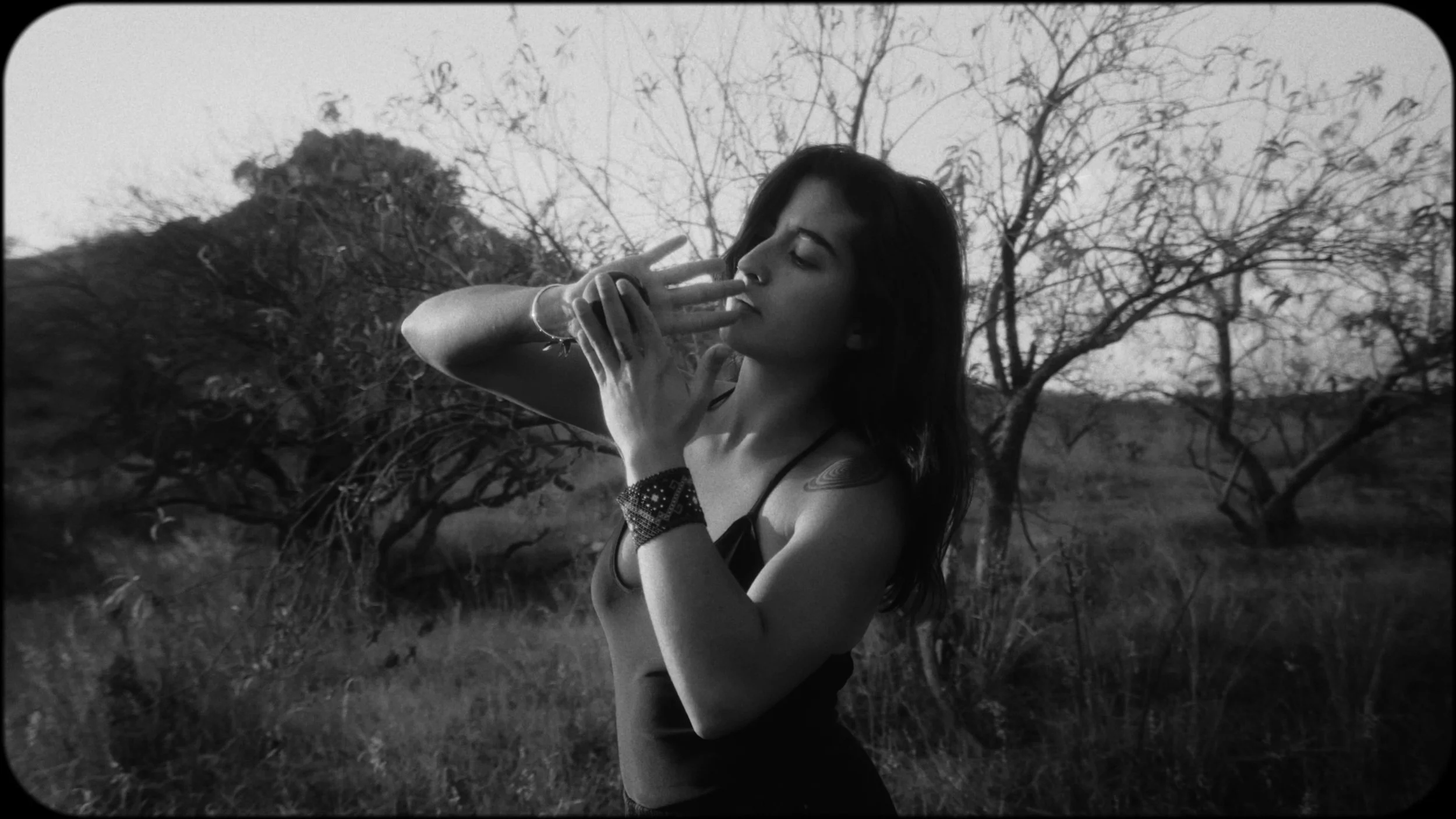 A woman with dark hair in a black dress and jewelry is standing outdoors in a field with bare trees in the background. She is holding two fingers to her lips in a pose, and the image is in black and white.