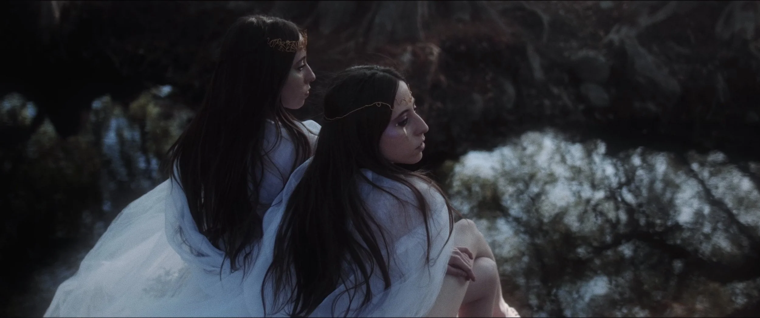 Two women with dark hair, wearing white garments, sitting by a rocky water body, gazing pensively into the distance during twilight.