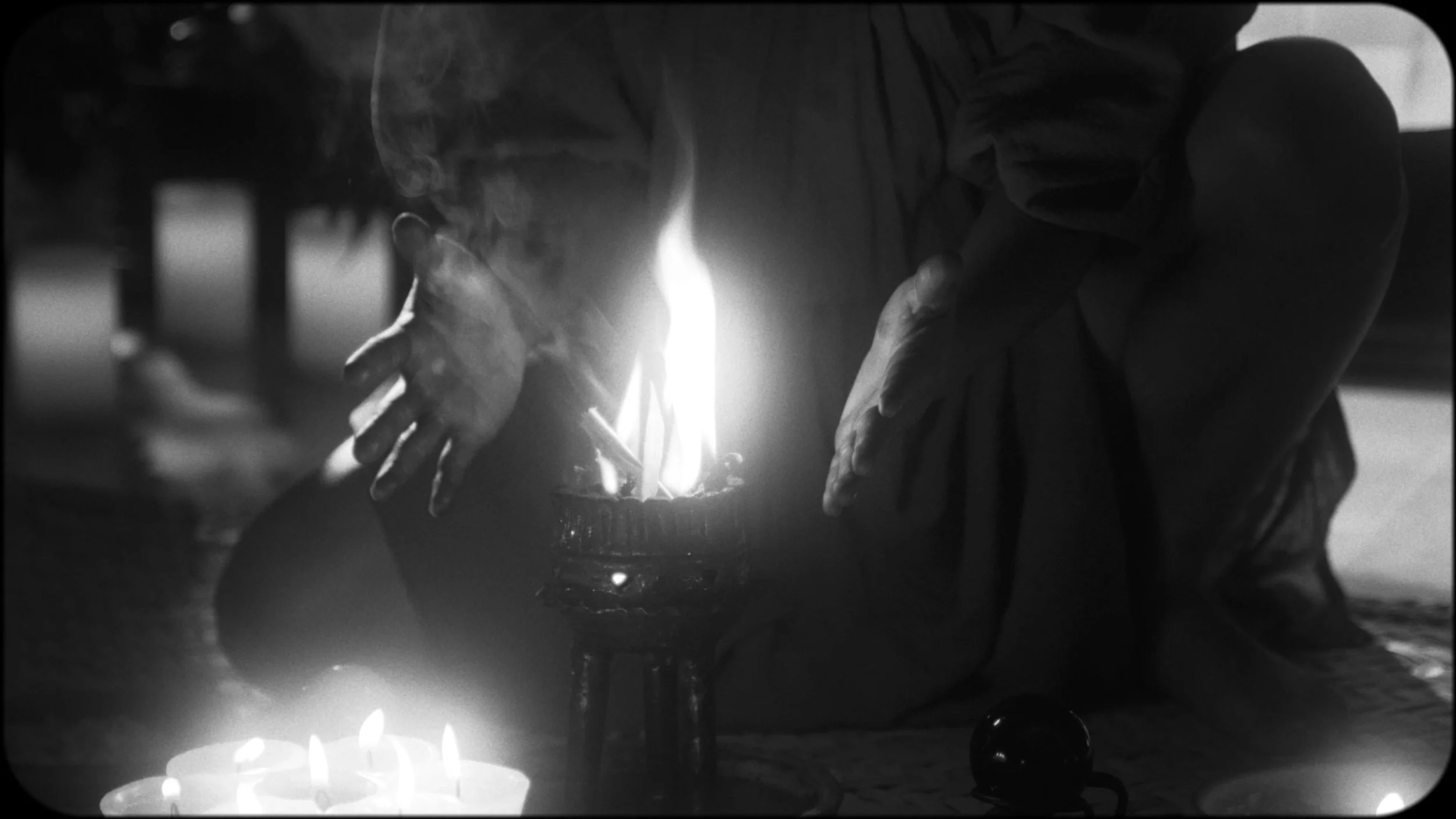 A person sitting on the ground, covering their face with their hands, illuminated by a small fire and surrounding candles in a dimly lit setting.
