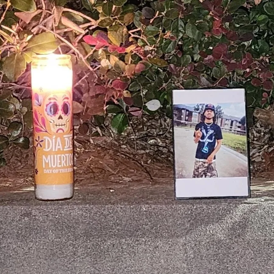 A lit candle with Día de Muertos decorations and a photograph of a young man standing outdoors, making a hand gesture, placed against a background of foliage.