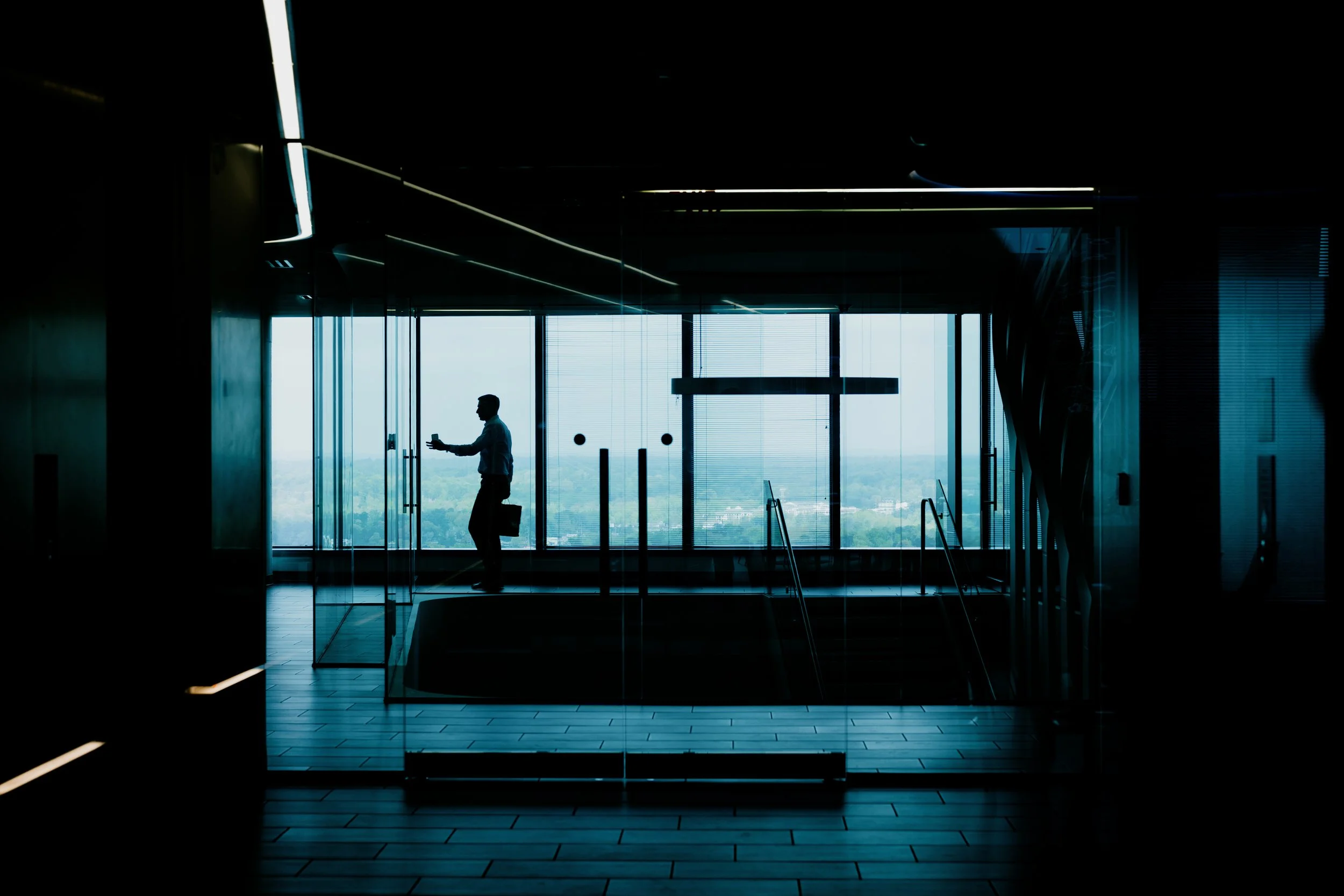 Silhouette of a man with a briefcase walking through a glass door in a modern office lobby with large windows showing a cityscape.