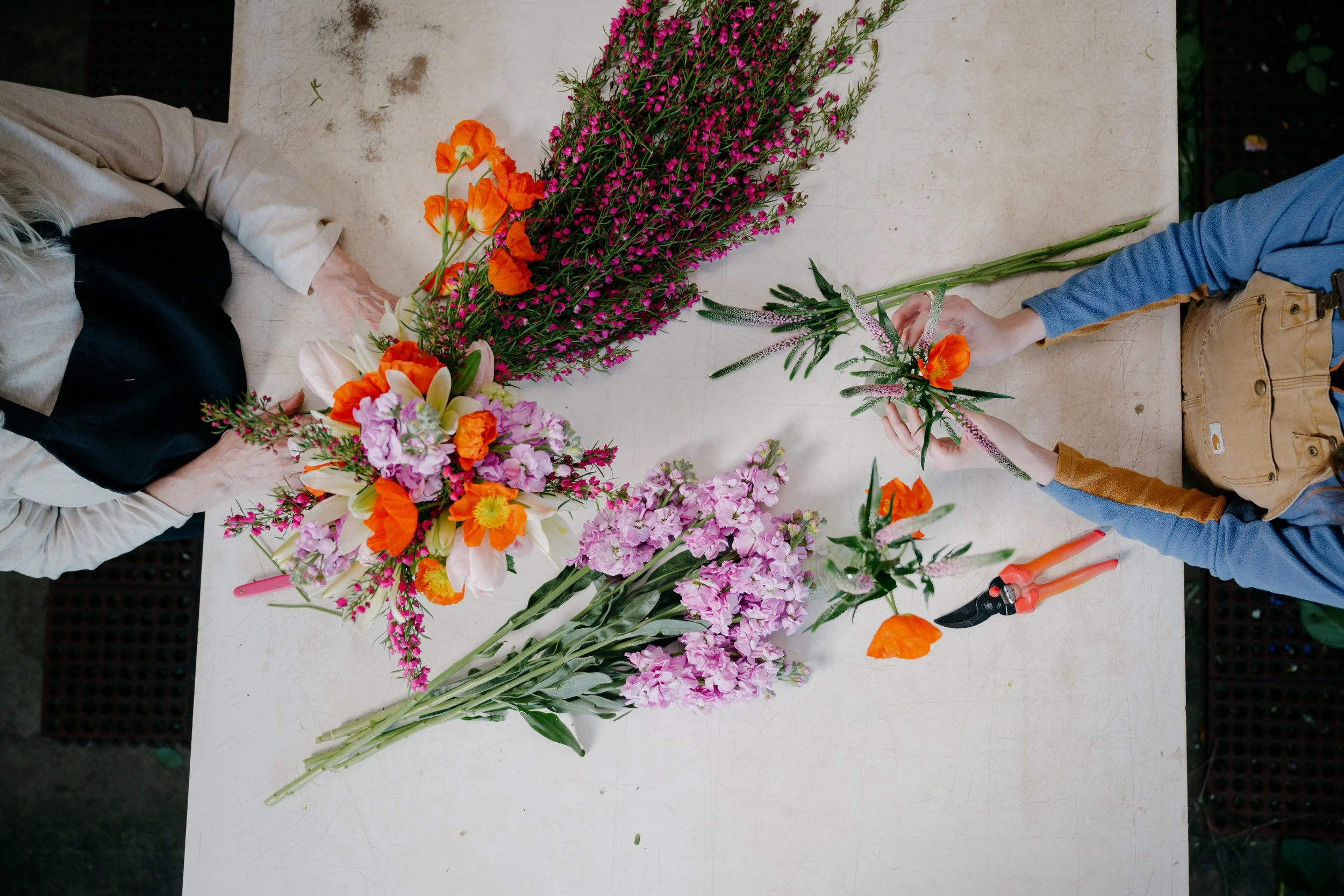 Two people arranging various colorful flowers on a white table. One person is wearing a beige jacket, and the other a blue shirt and tan pants. Flowers include orange poppies, pink and white lilies, and purple and pink small blooms.