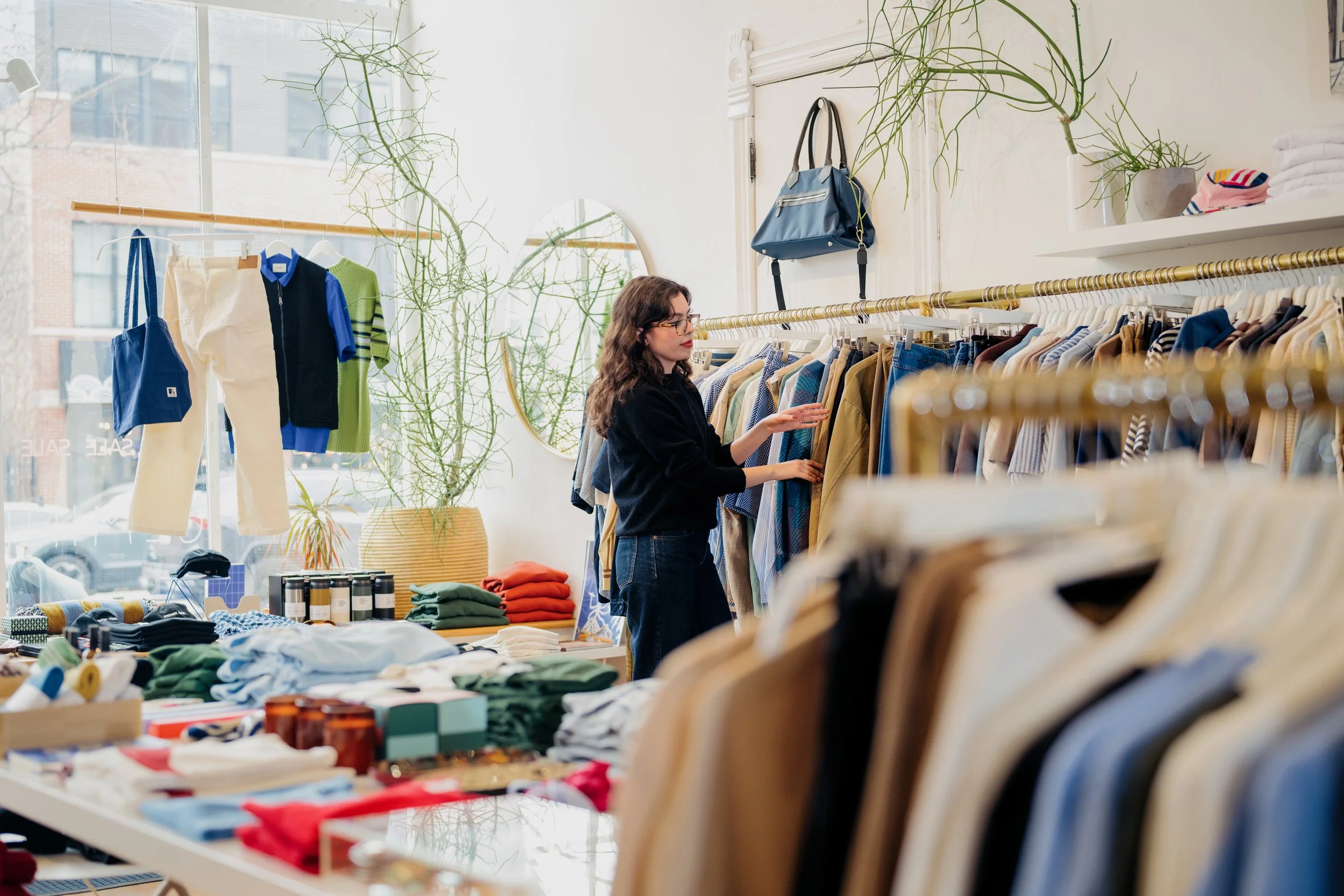 A woman shopping in a clothing store with racks of clothes, folded garments, and accessories. Large windows with city buildings outside, and indoor plants visible.