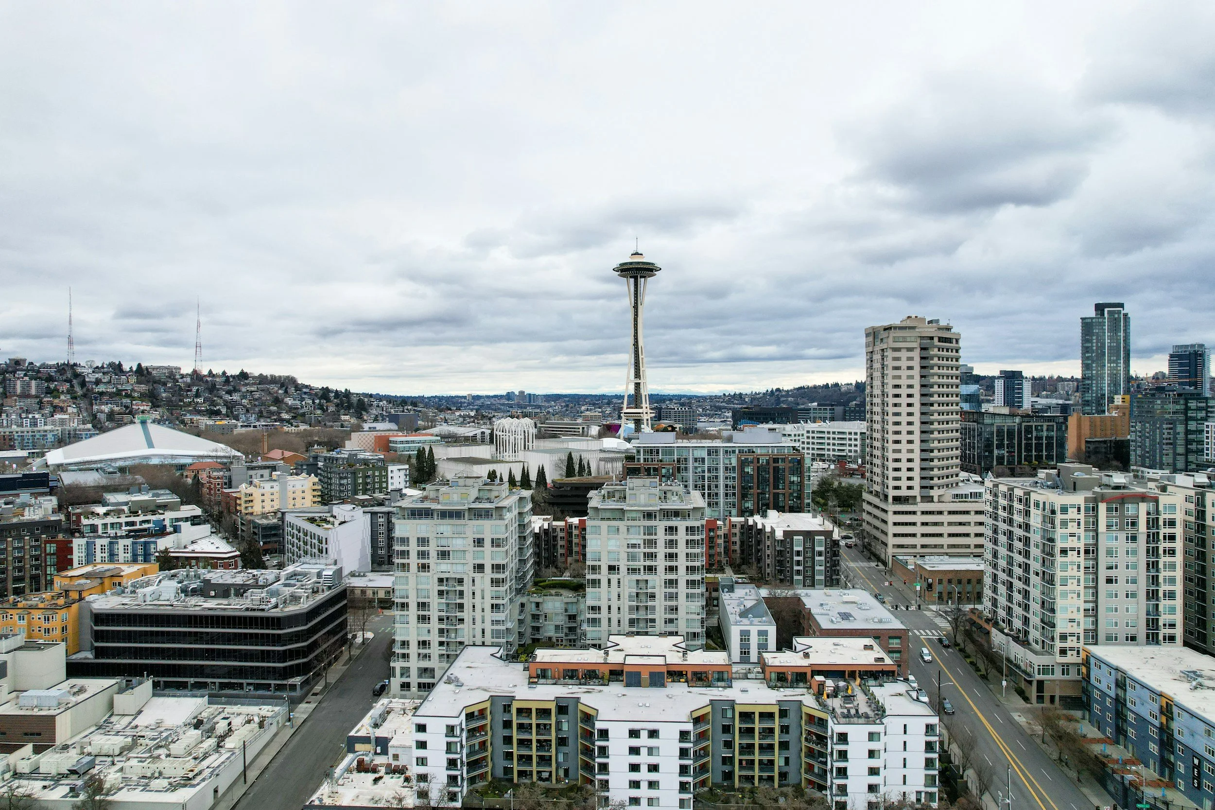 A city skyline featuring the Space Needle in Seattle under cloudy skies, surrounded by modern high-rise buildings and residential apartments.