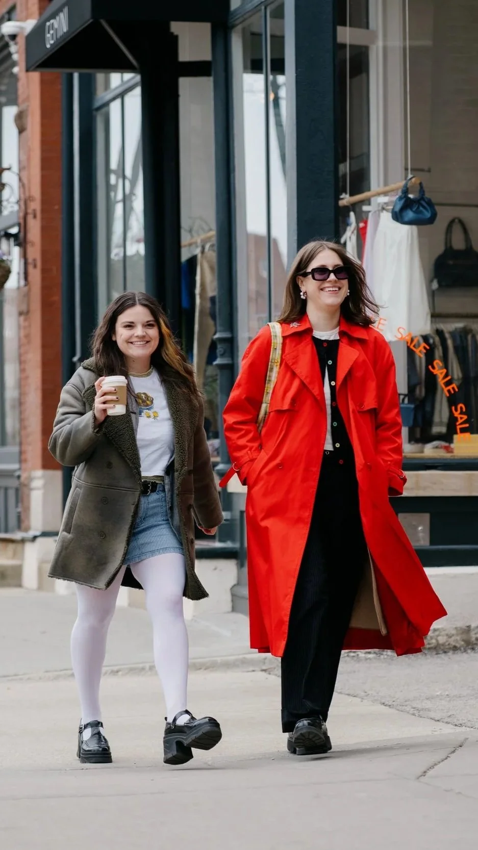 Two women walking outside a store, smiling, one holding a coffee cup. One wearing a gray coat, white tights, and platform shoes; the other in a red trench coat with sunglasses and a plaid bag.