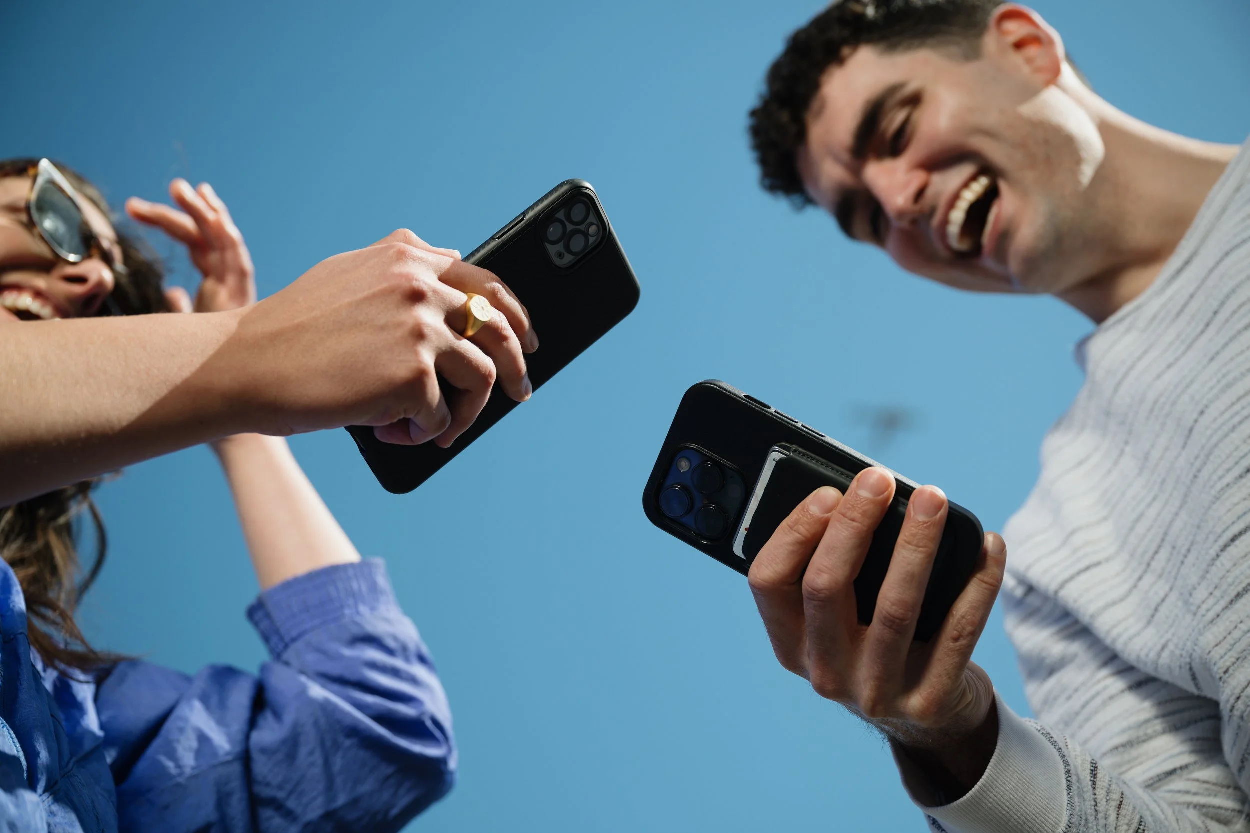 Two people, a woman and a man, are smiling and looking at their smartphones with the blue sky in the background. The woman is wearing glasses and a blue jacket, and the man is wearing a light gray shirt.