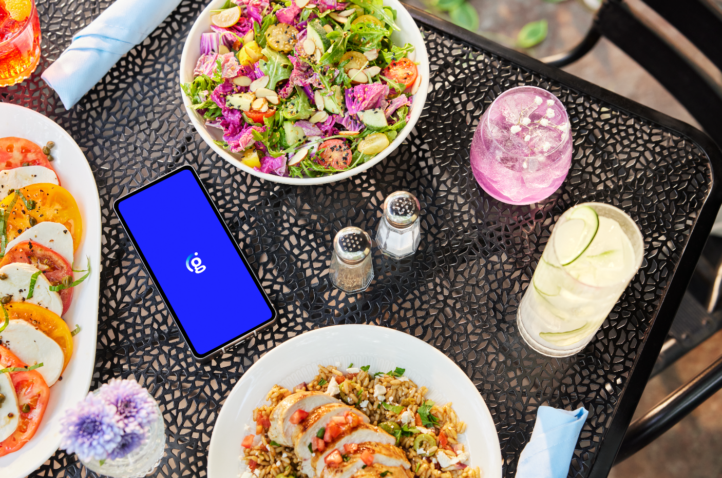 Outdoor table with a colorful salad, sliced tomato and mozzarella, rice dish with sliced chicken, two drinks with cucumber slices, salt and pepper shakers, a smartphone, and a small purple flower vase. Restaurant and lifestyle photography.