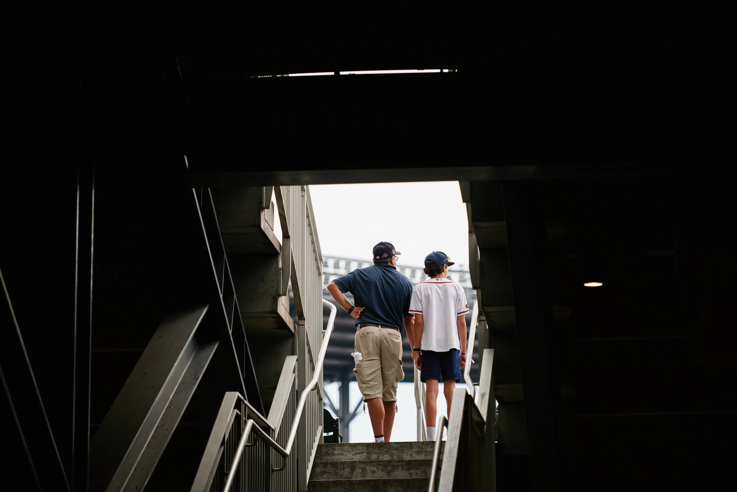 Two men walking up a set of stairs underneath a dark overhang, with bright light behind them, outside a stadium or event venue.