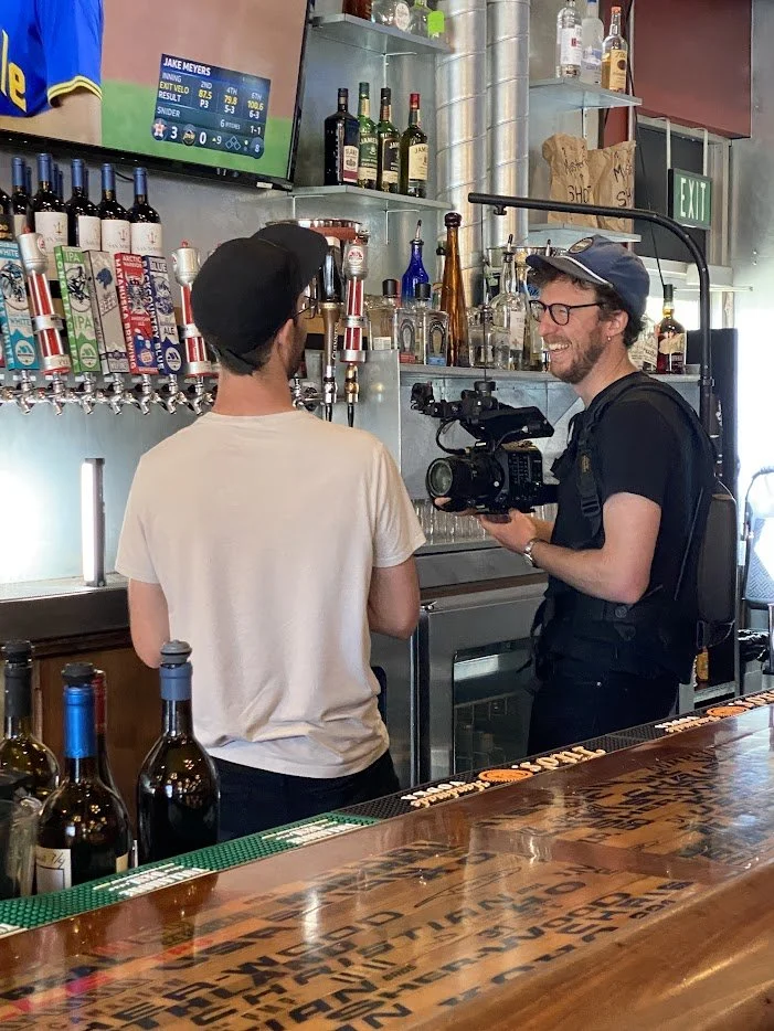 A man in a black shirt and glasses being interviewed by a cameraman at a bar with bottles and taps for drinks in the background.