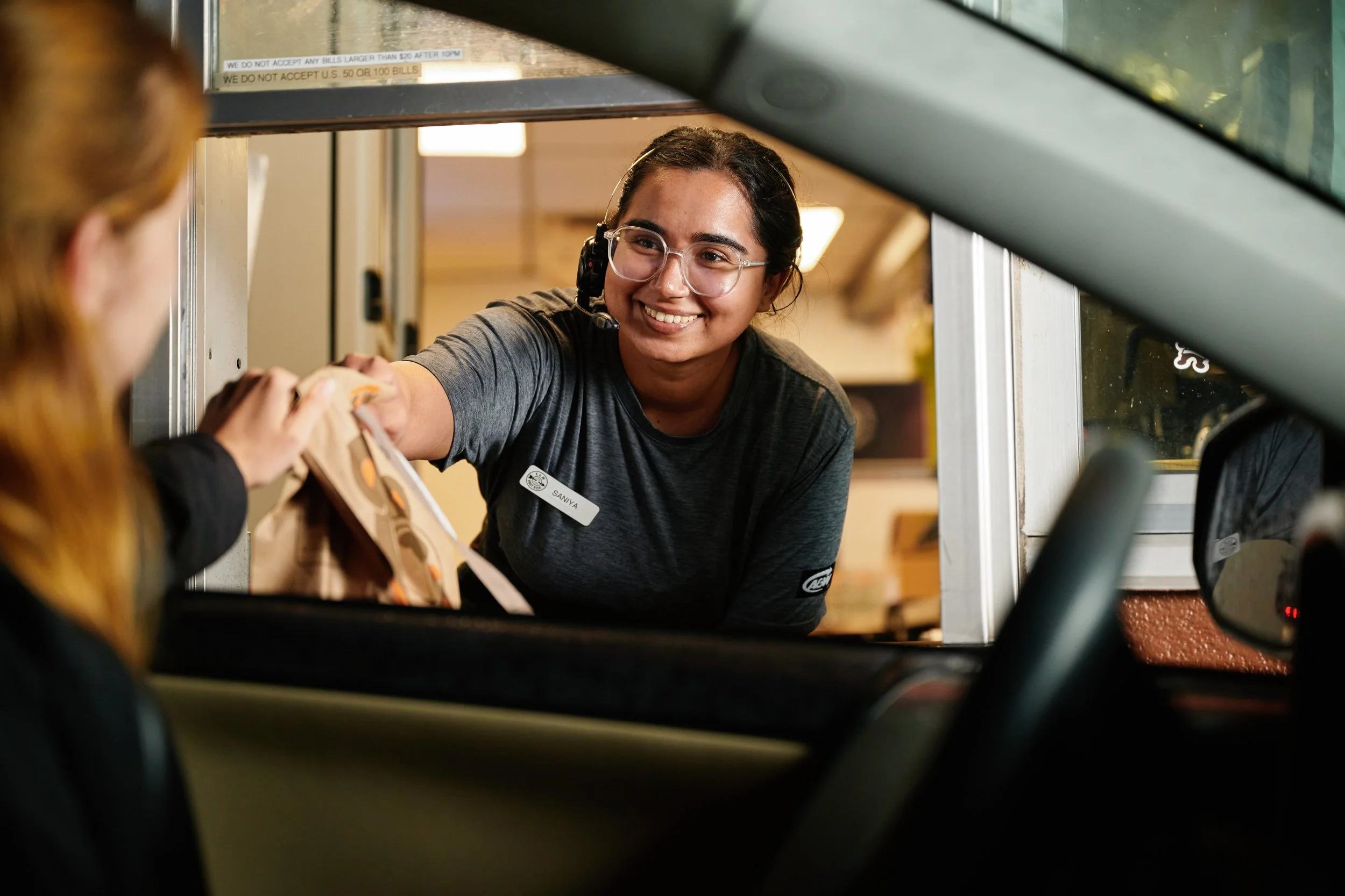 A costumer at a fast food or drive-thru window handing money or an order to a smiling female employee wearing glasses and a headset at a restaurant or concession stand. Quick serve restaurant photography. 