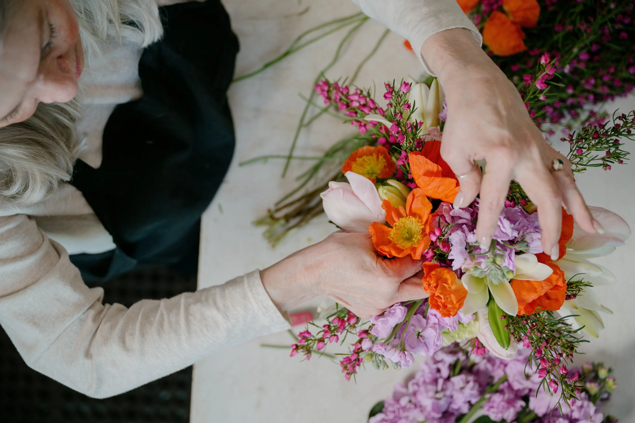 A person with blonde hair arranging a colorful bouquet of flowers, including orange, pink, and purple blossoms, on a white surface.