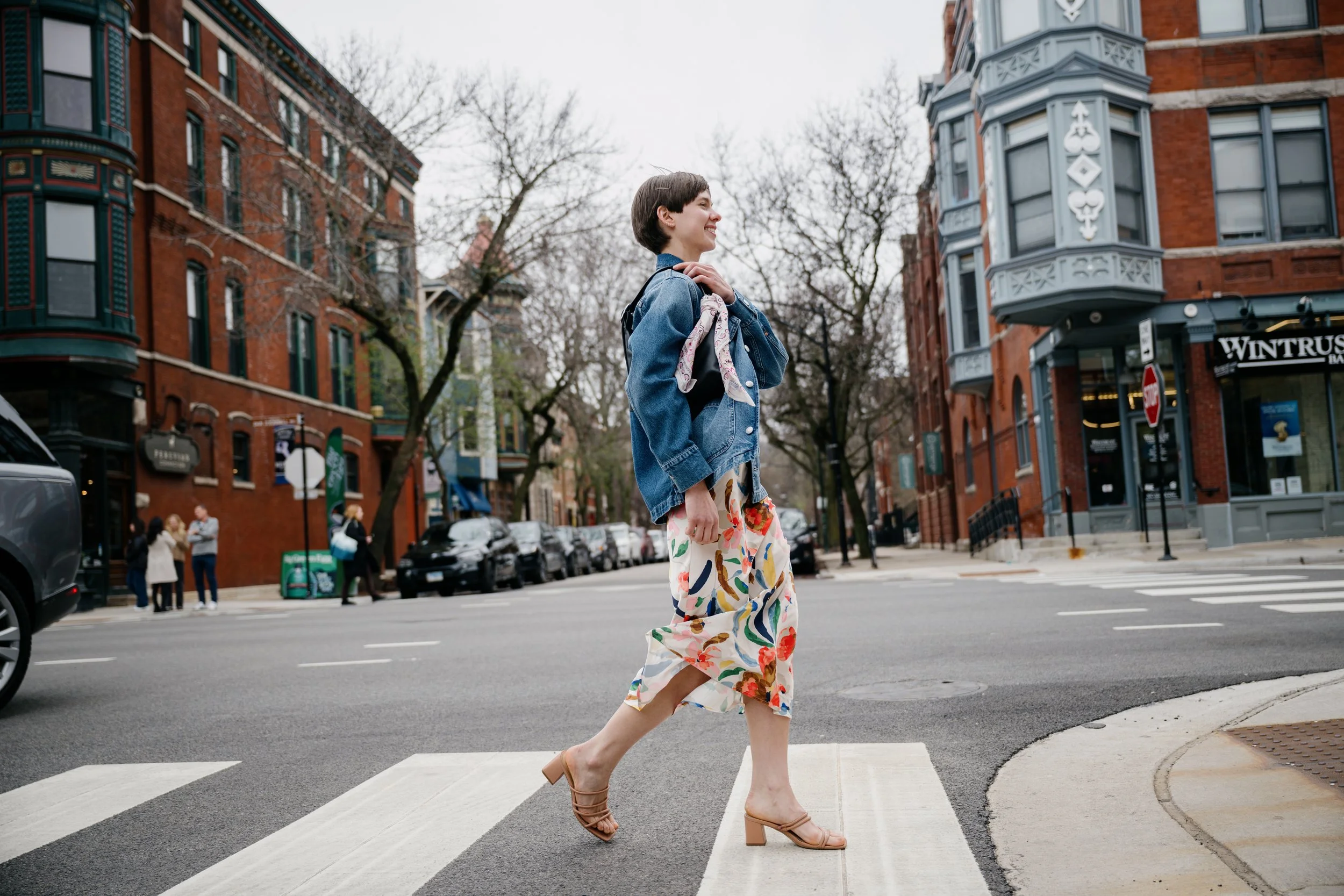 A woman in a denim jacket and colorful floral pants is crossing the street at a crosswalk, smiling, with her hand on her chest, in an urban neighborhood with brick buildings and leafless trees.