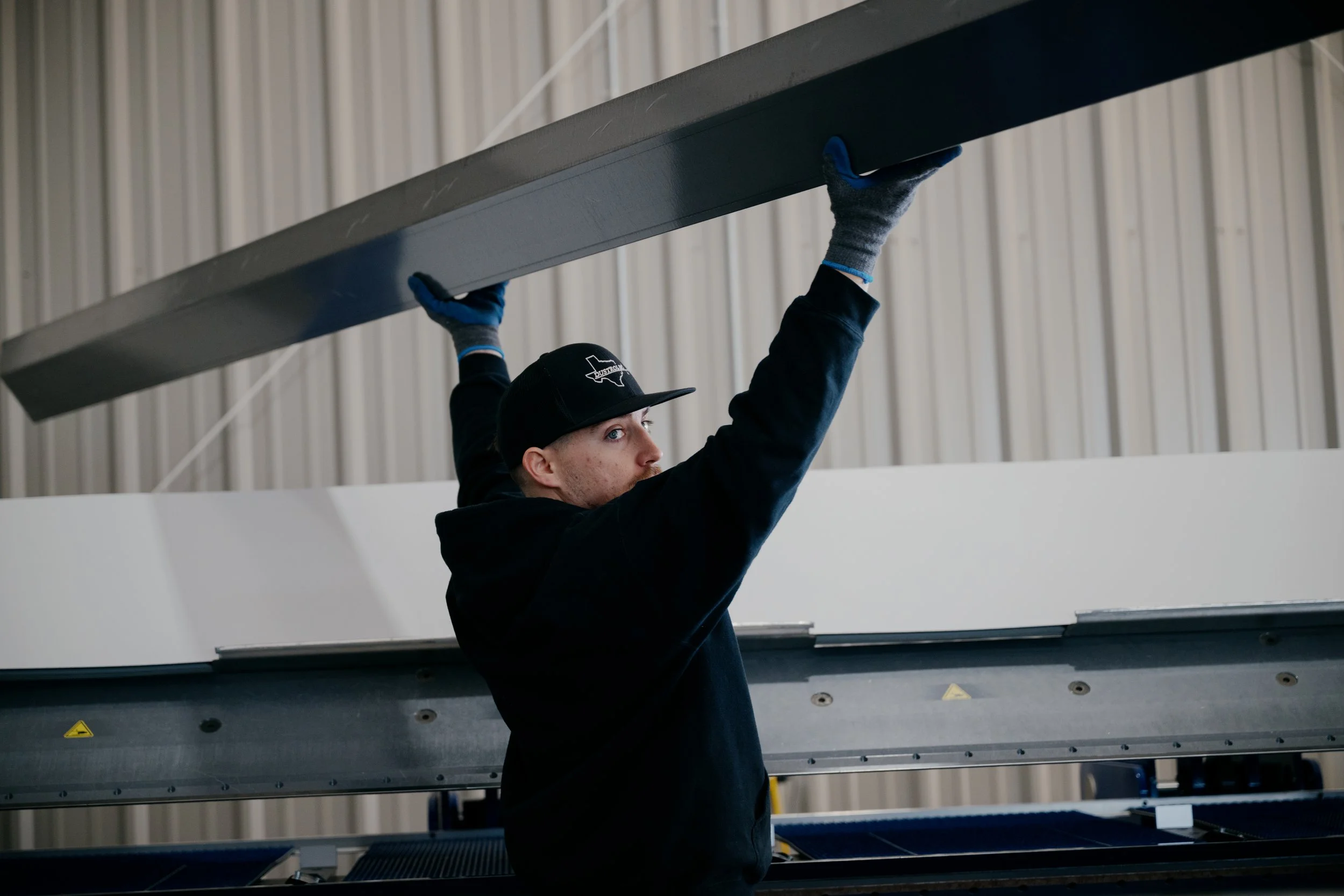 A person wearing a black cap, black hoodie, and gray gloves is lifting or installing a large gray panel or piece of equipment in an industrial or warehouse setting, with a high ceiling and metal siding in the background. Construction photography. 