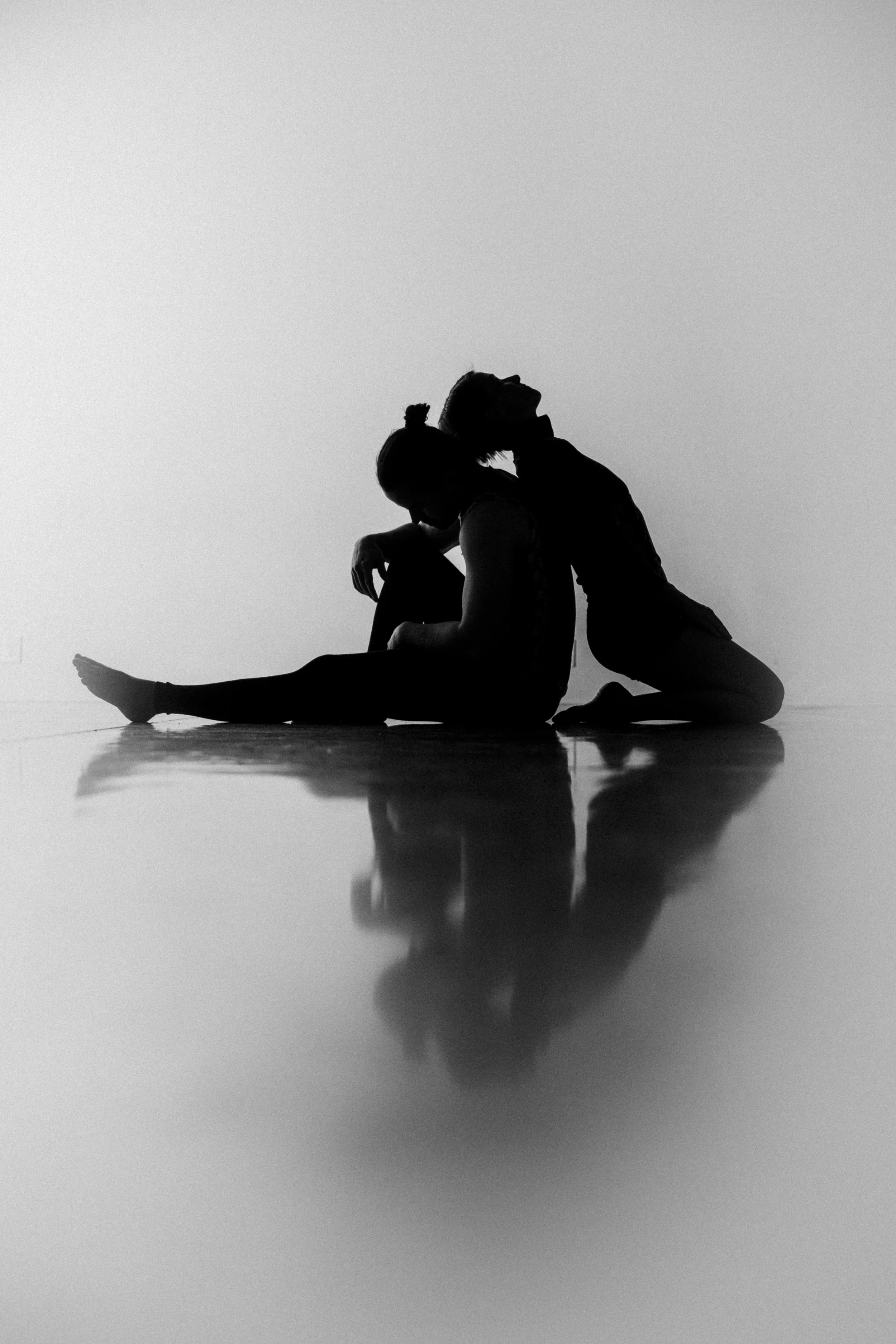 Silhouette of two people sitting back-to-back on the floor with a reflection visible below, in front of a plain background.
