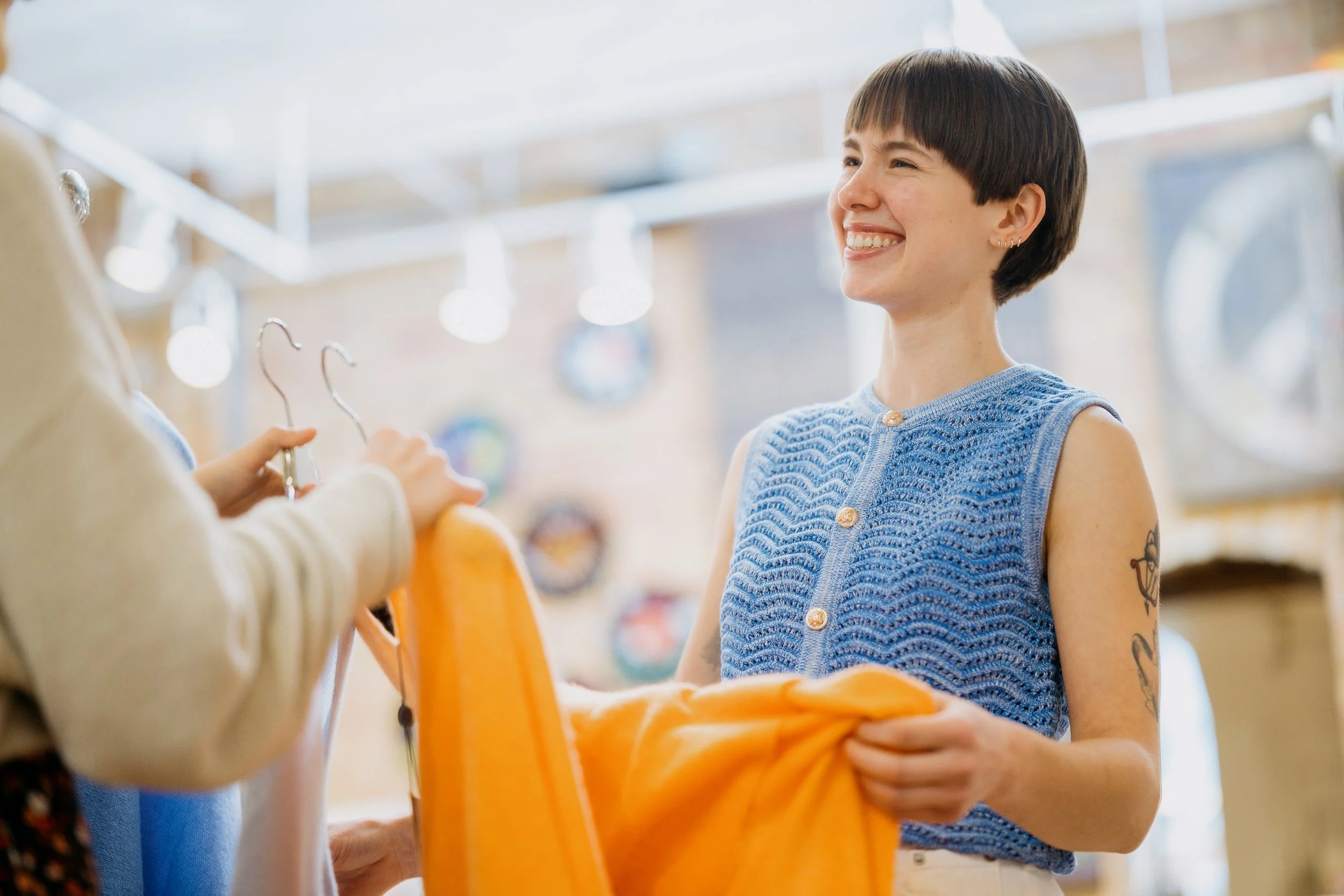 A woman with short dark hair and tattoos on her arm smiling while shopping for jackets in a store. Retail and lifestyle photography featuring warm, bright expressions and a light and airy color grade. 