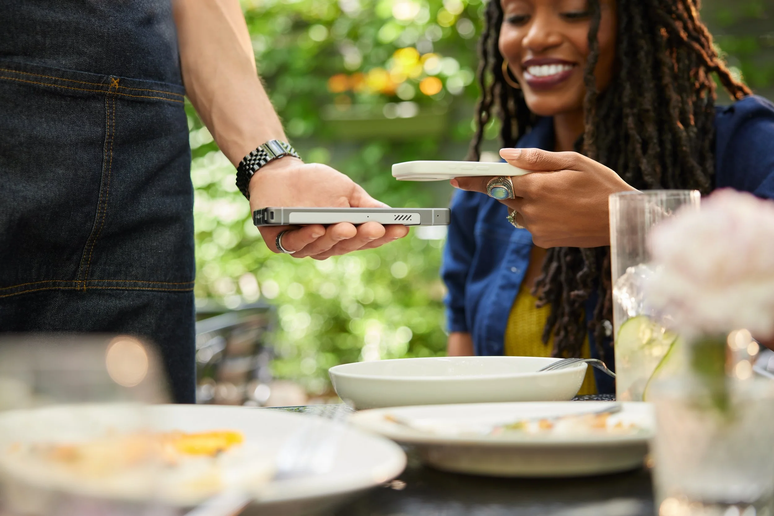 A woman with long dreadlocks smiling while holding a plate and a phone, standing near a table with food outdoors with blurred greenery in the background.